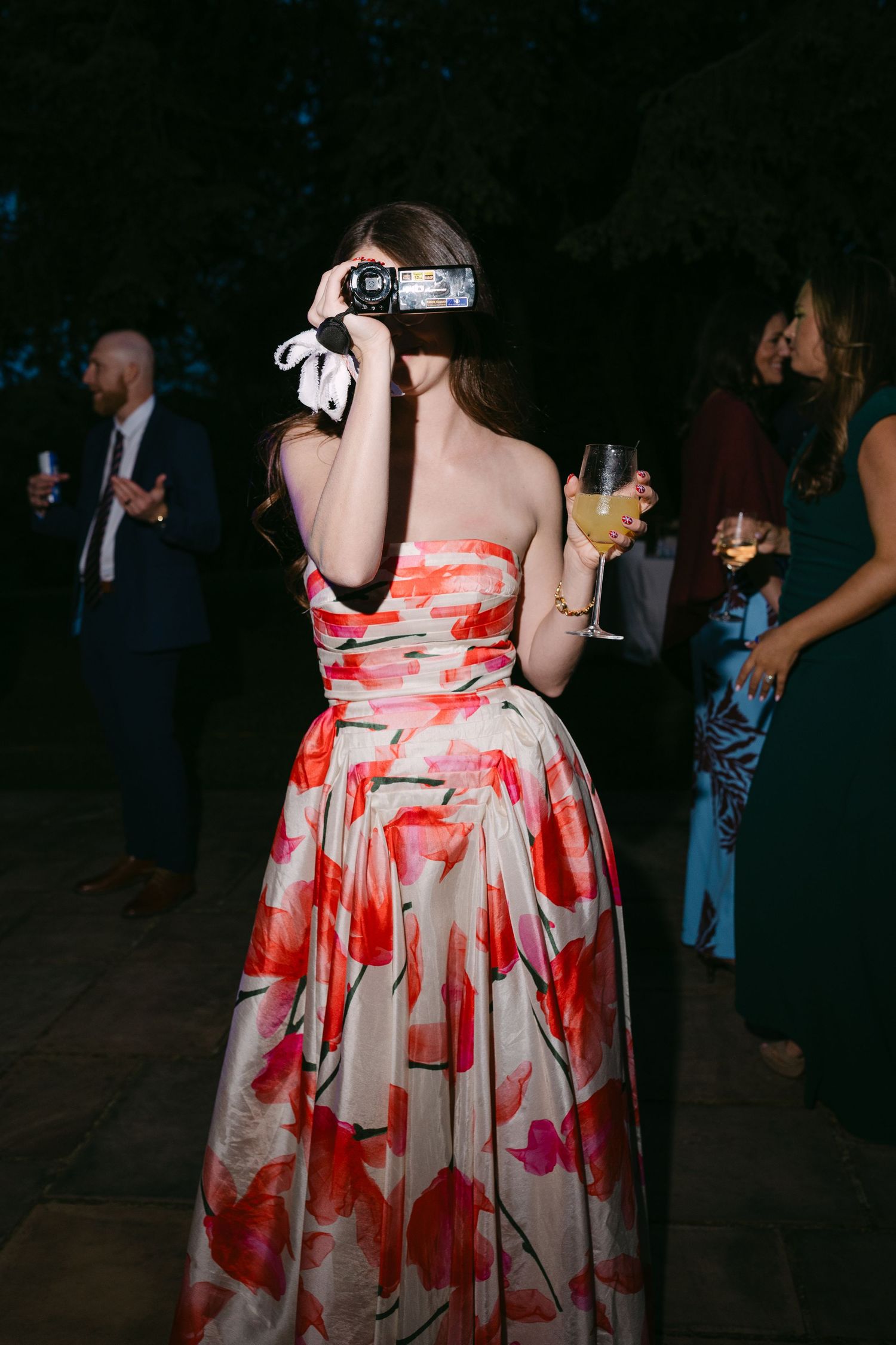 Wedding guest in floral strapless gown filming with camcorder while holding drink at reception.