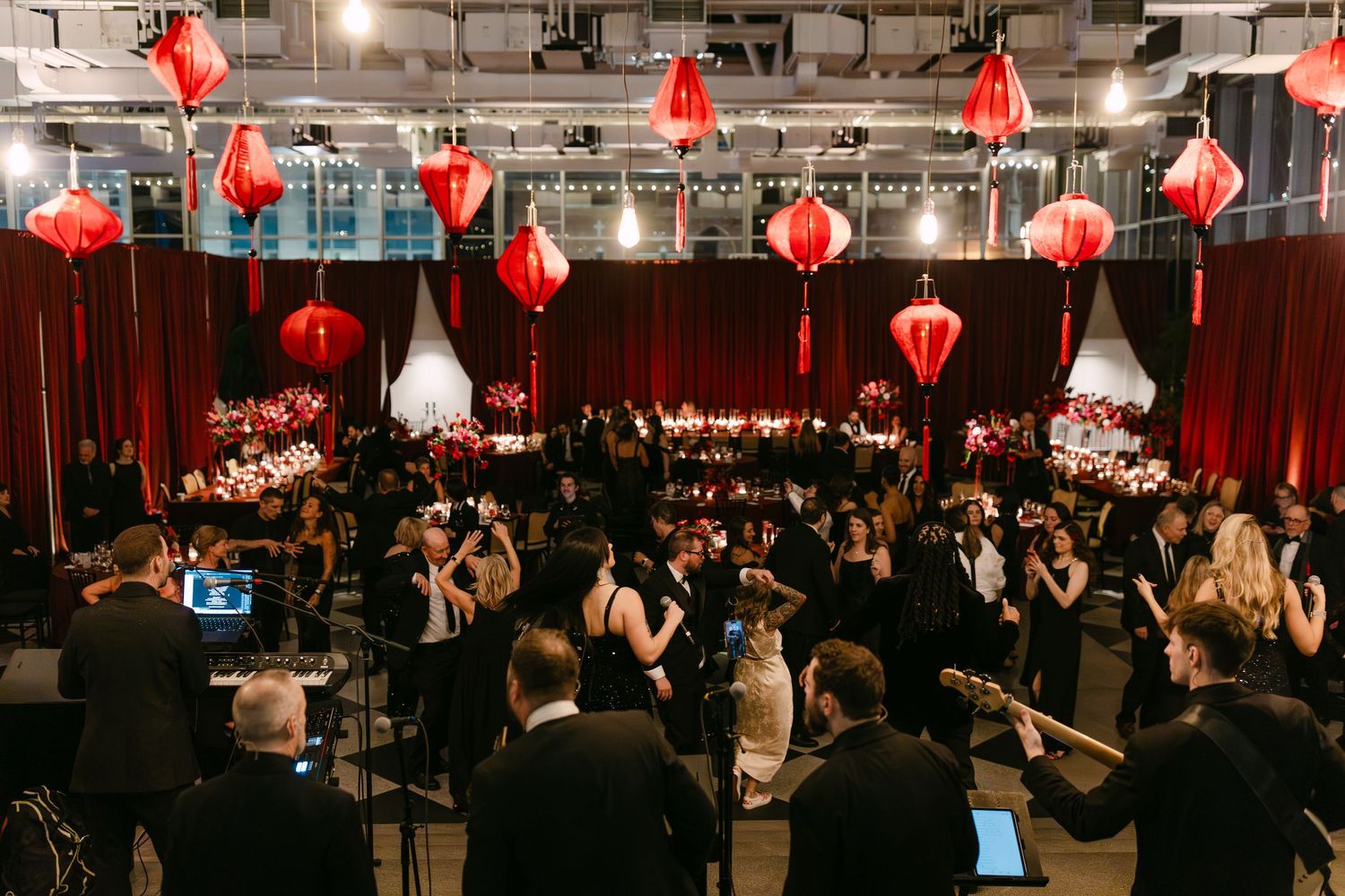 Elegant wedding reception hall decorated with red Chinese lanterns, red draping, and floral centerpieces with live band.