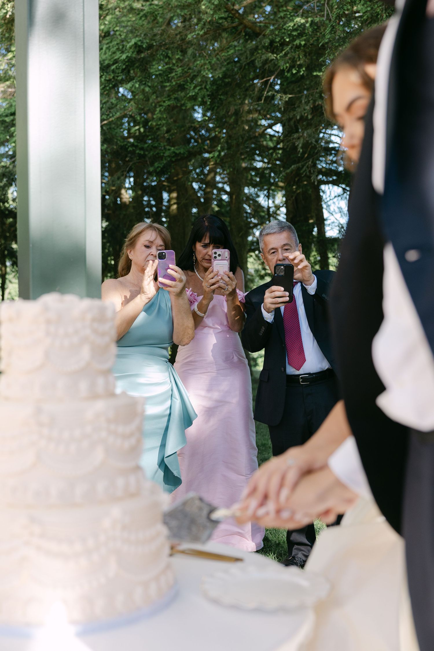 Wedding guests photograph bride and groom cutting white tiered cake outdoors among green trees.