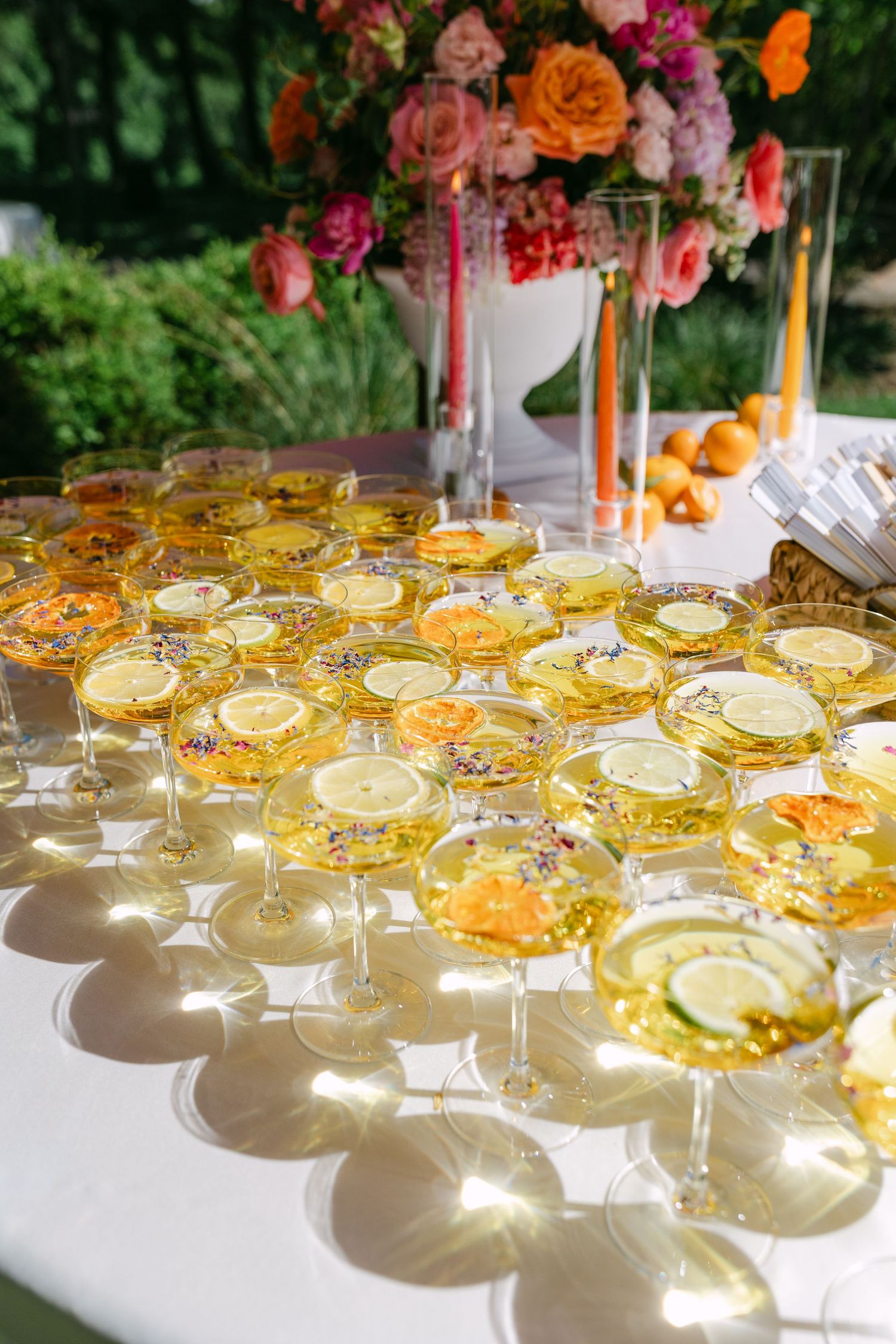 Elegant outdoor wedding cocktail hour with champagne coupes garnished with lemon slices and edible flowers on a white tablecloth.
