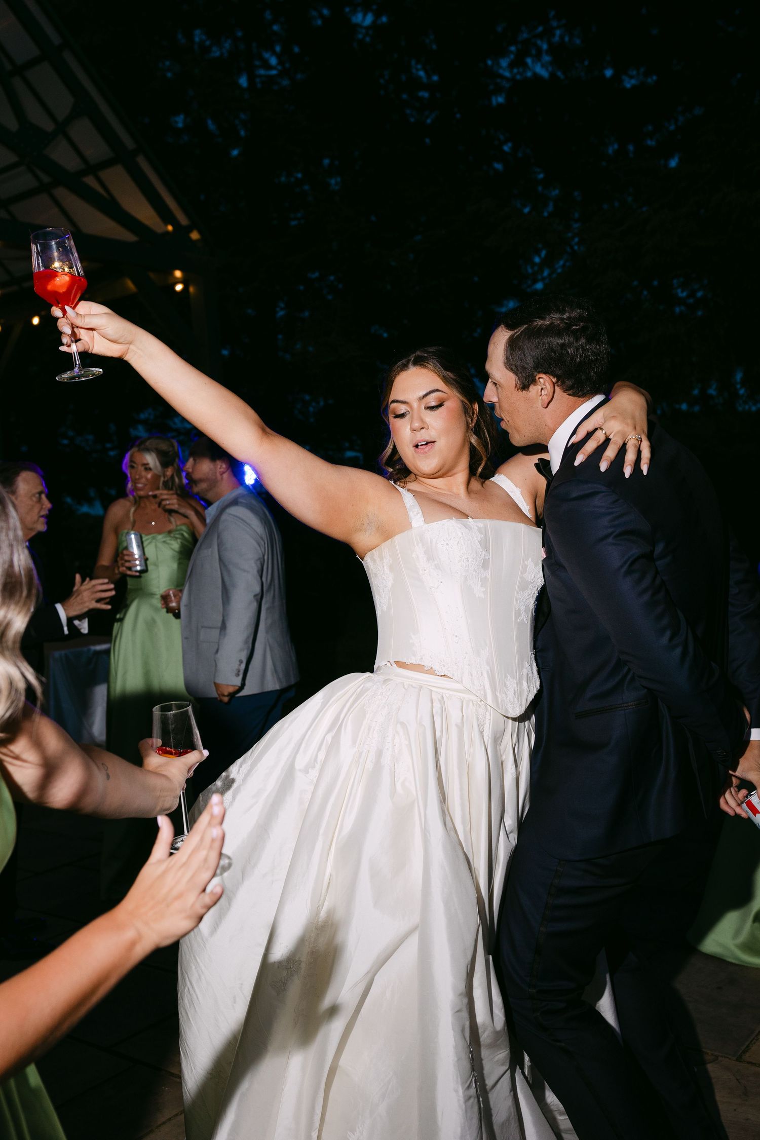 Bride in white gown raises a red cocktail while dancing with groom at an outdoor evening wedding reception.