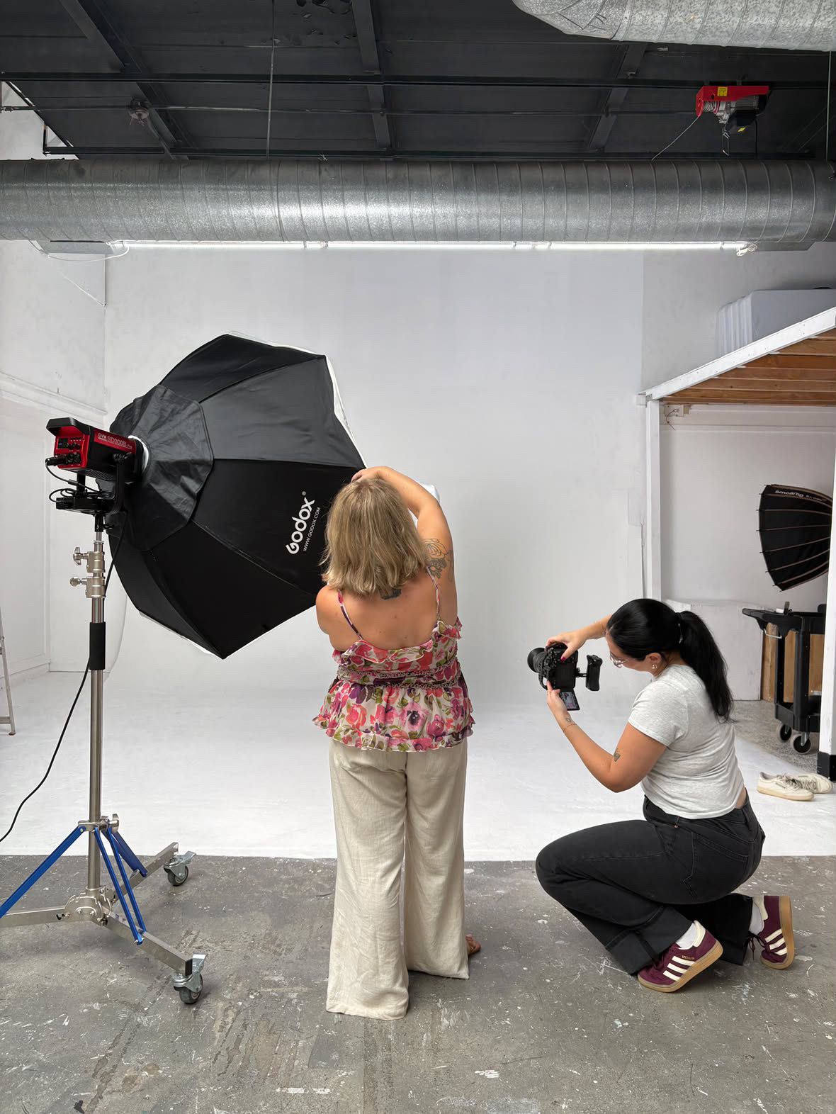 Sarah Puliatti Thomas crouching to photograph blonde woman in floral top against white backdrop in photography studio.