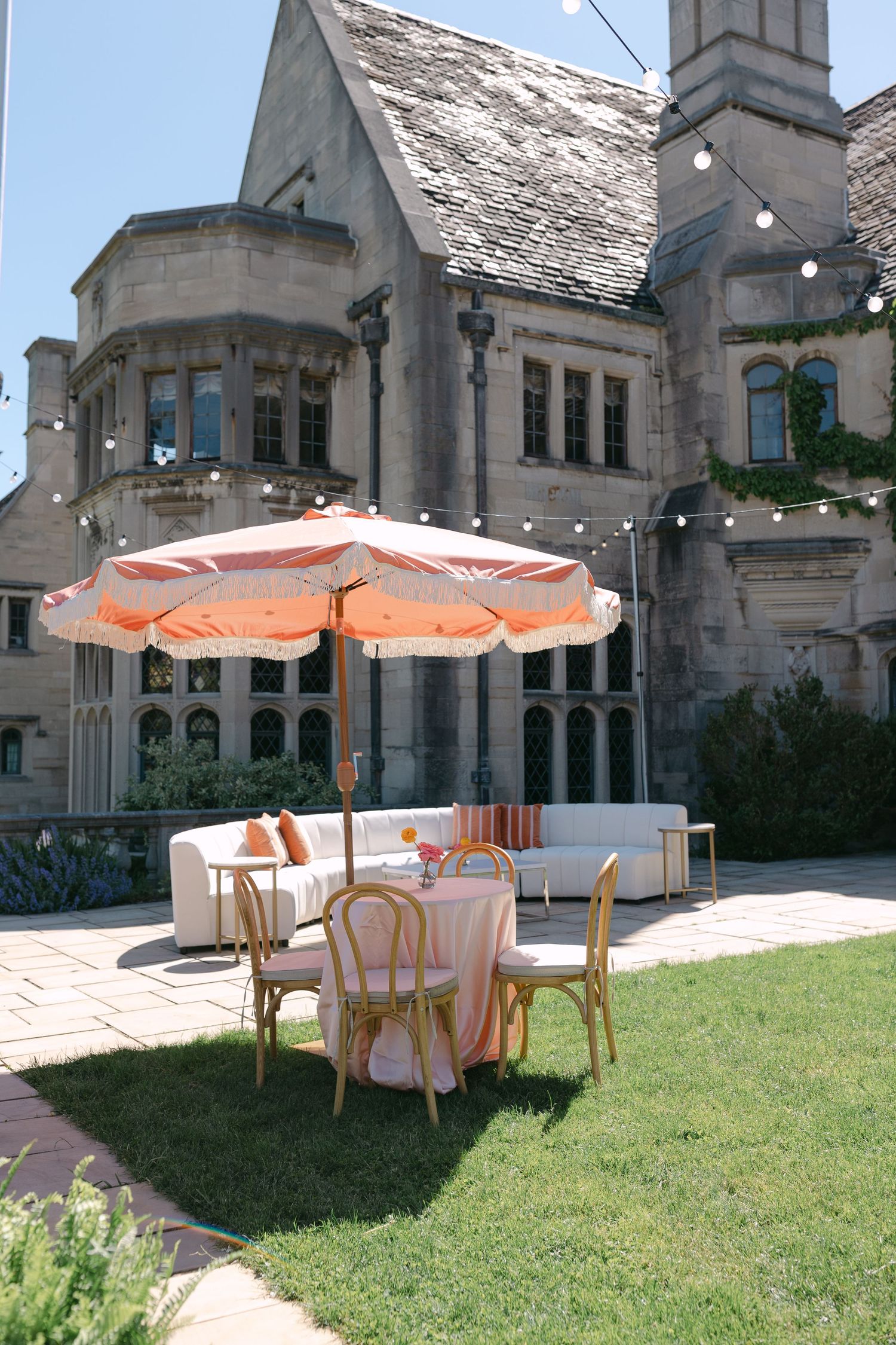 Elegant outdoor wedding setup with orange striped umbrella and seating area in front of stone manor.