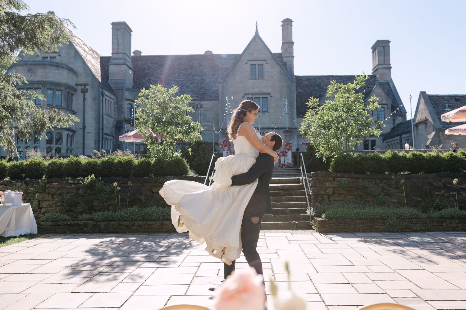 Groom lifts bride in white gown outdoors in front of elegant stone mansion wedding venue.