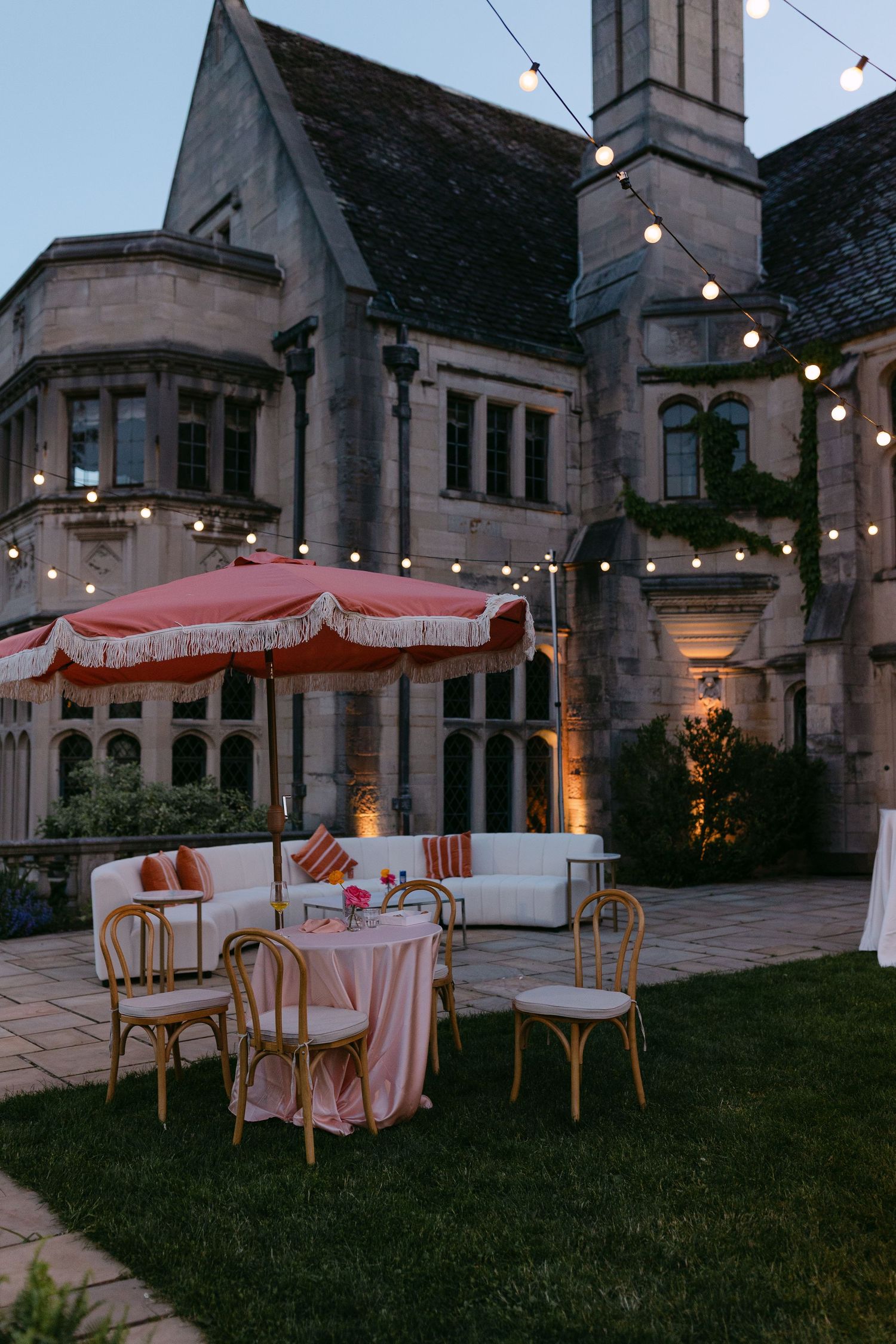 Romantic outdoor wedding lounge with pink fringed umbrella, white sofas, and string lights against a stone manor at dusk.