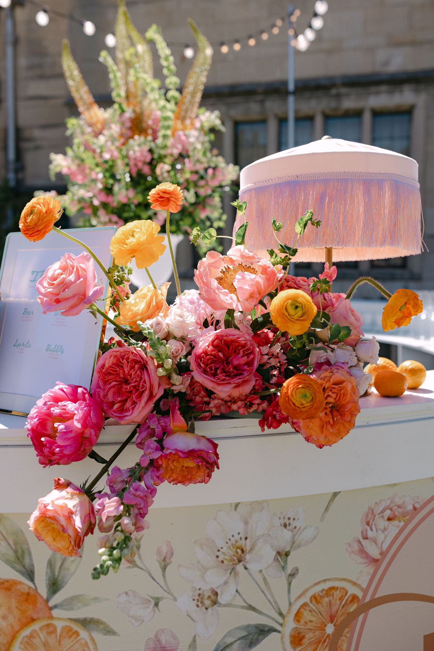 Vibrant pink, orange, and coral floral arrangement with fringed lamp on floral-printed wedding welcome table.