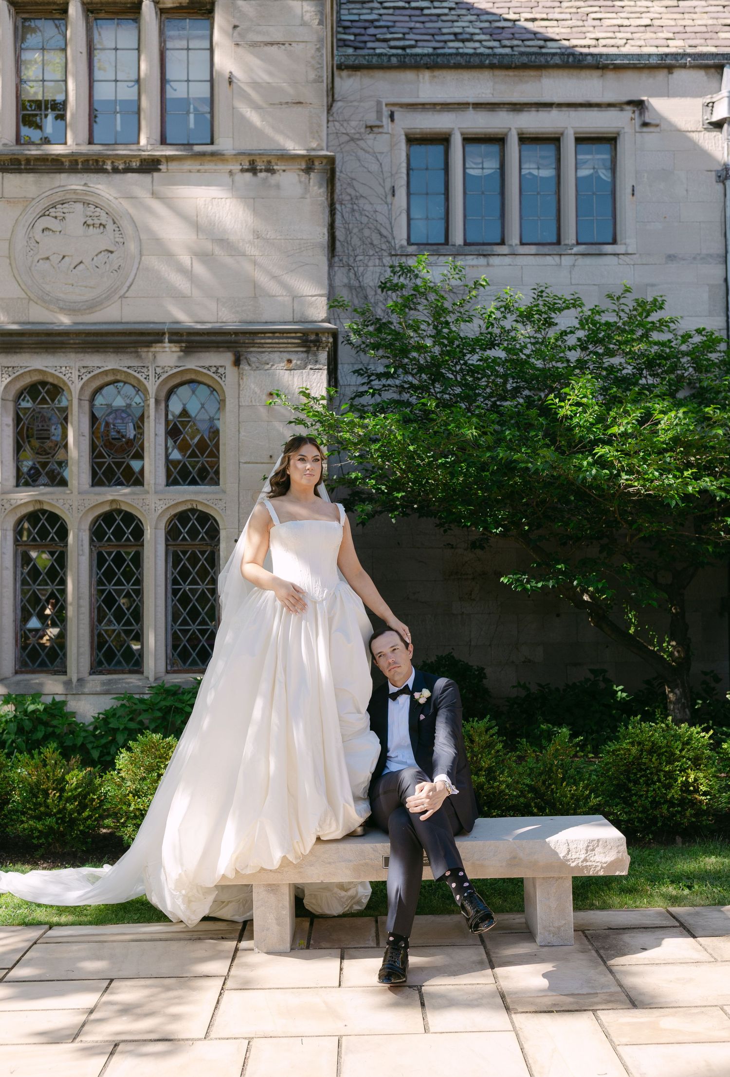 Bride in a white ball gown and groom in a tuxedo pose by a stone bench at a historic estate.
