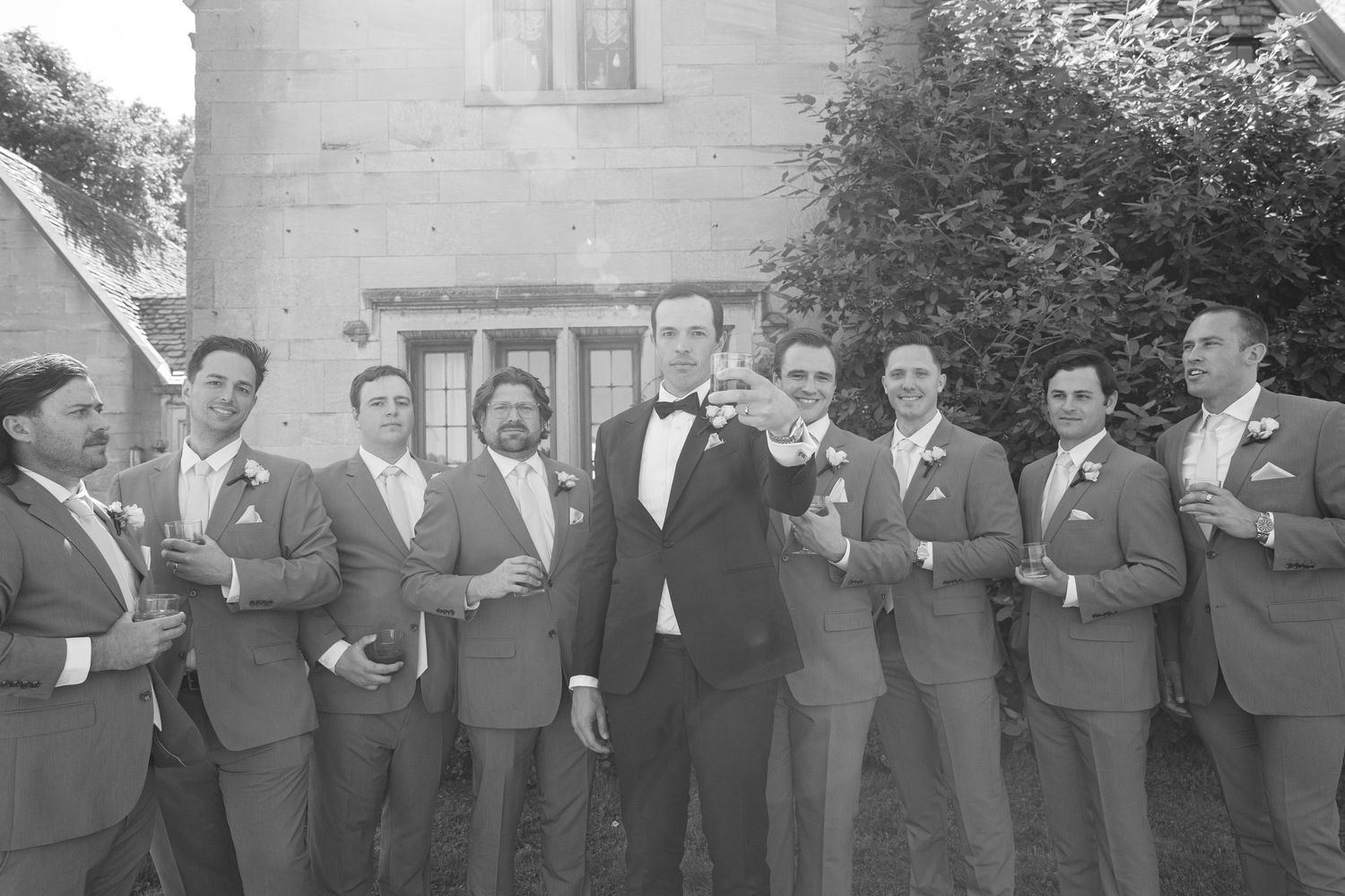 Black and white photo of groom in tuxedo with groomsmen in suits raising glasses outside stone building.