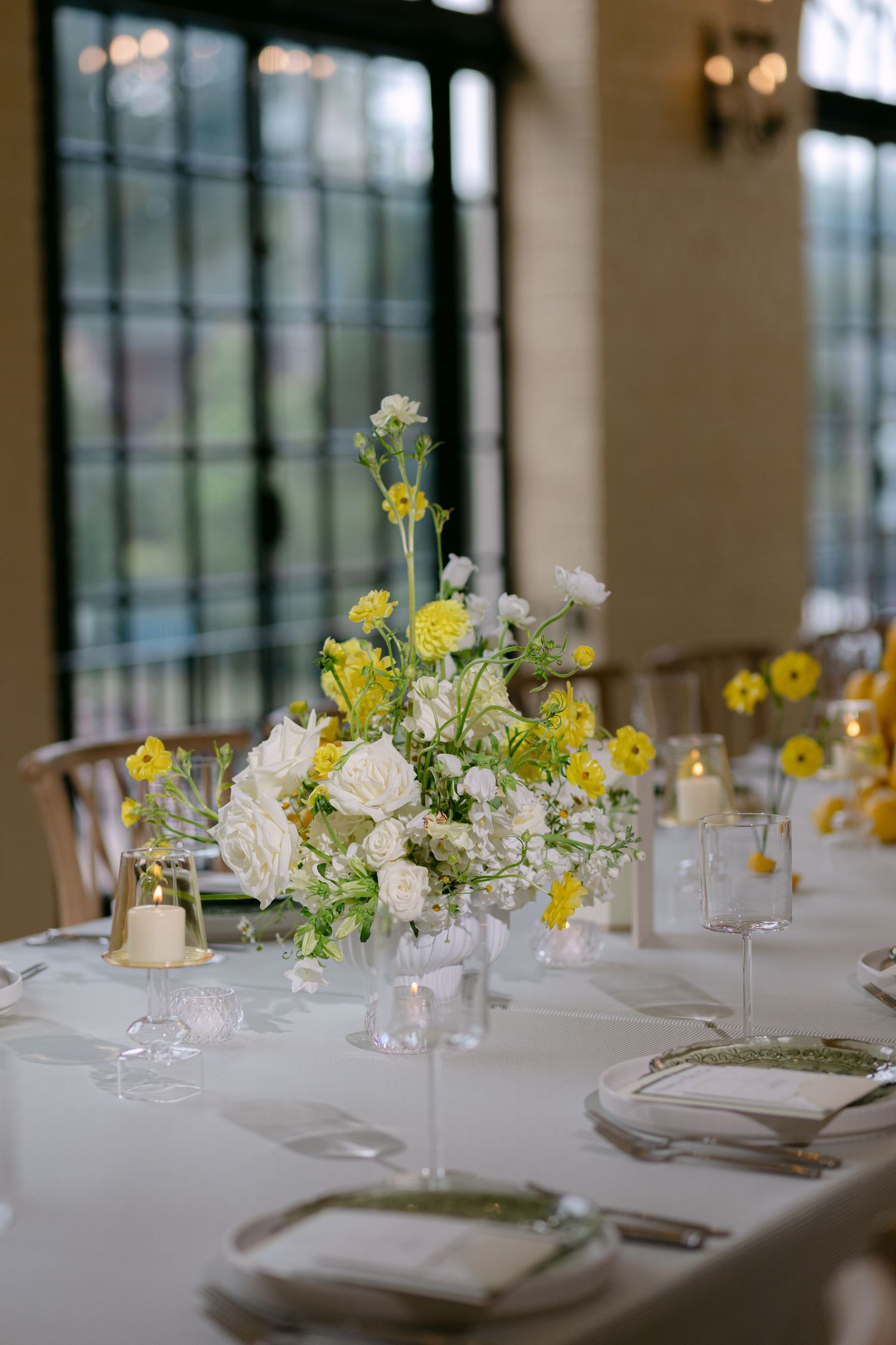 Yellow and white floral centerpiece on a white-draped wedding reception table near large windows.