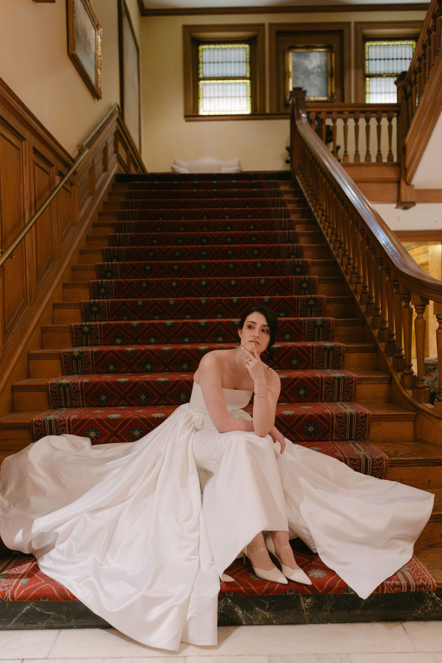 Bride in a white ball gown sitting gracefully on a grand staircase with red carpet and wooden railings.