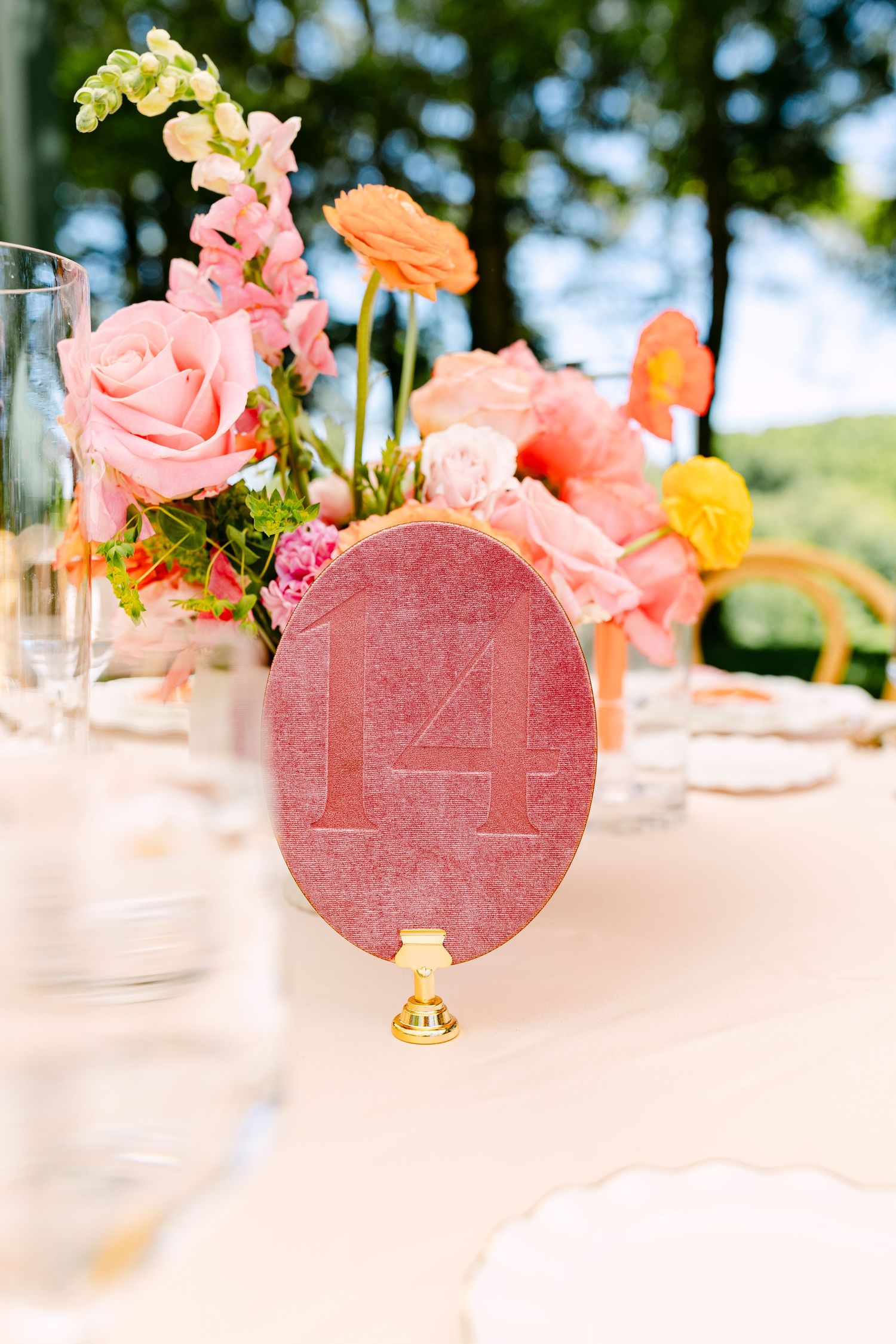 Pink velvet oval table number 14 on gold stand with colorful floral centerpiece at wedding reception.