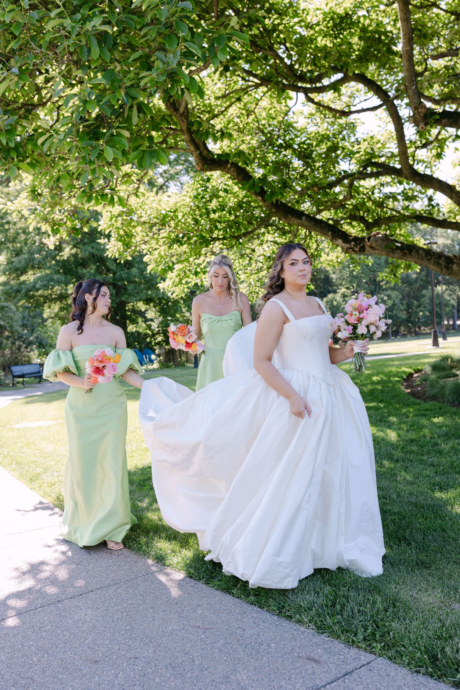 Bride in white ball gown walking with bridesmaids in sage green dresses carrying colorful bouquets outdoors.