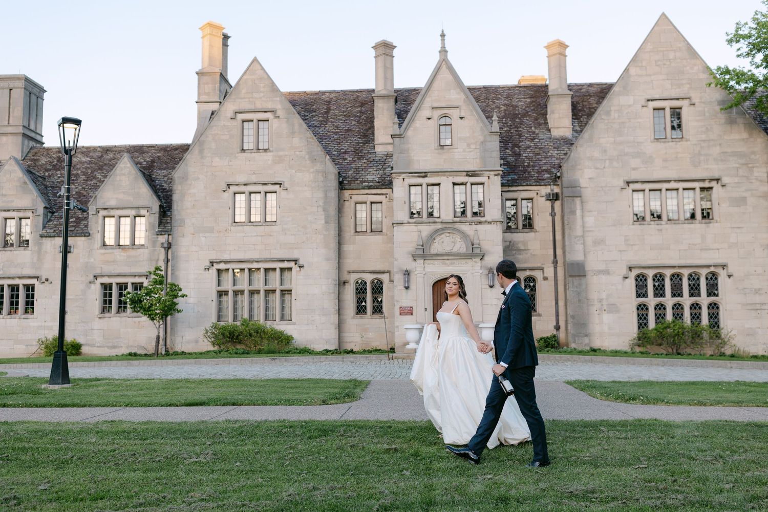 Bride and groom walking hand in hand on green lawn in front of grand stone manor at dusk.