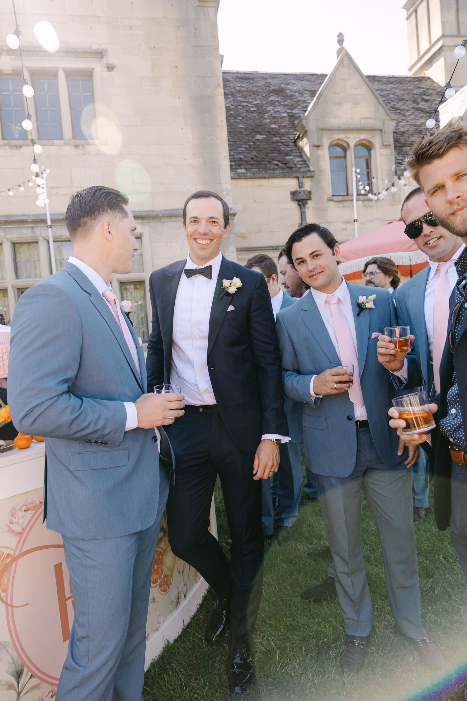 Groom in black tuxedo with bow tie socializing with groomsmen in blue suits during outdoor wedding cocktail hour.