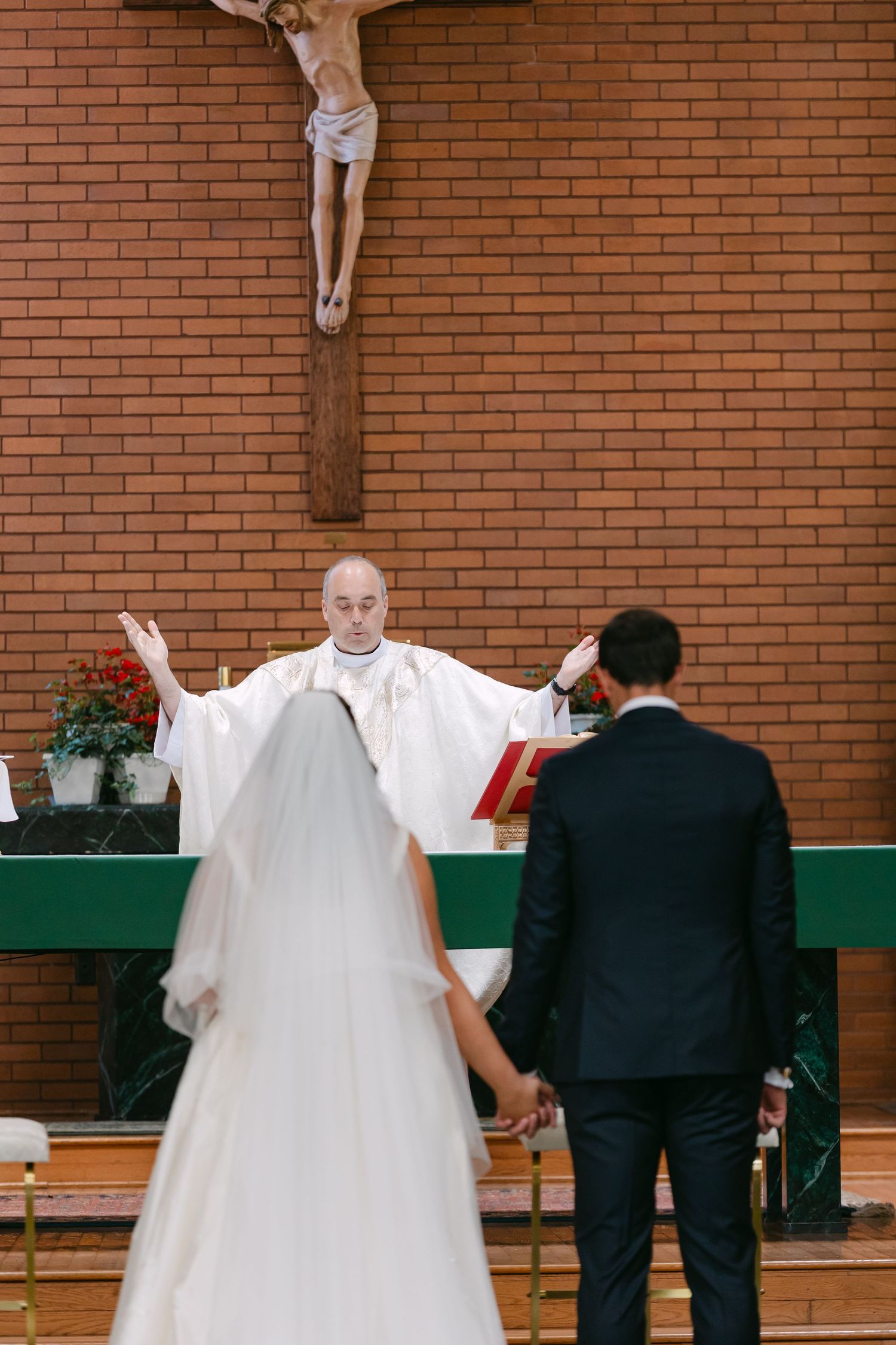 A priest with arms raised blesses a bride and groom holding hands at a Catholic wedding ceremony altar.