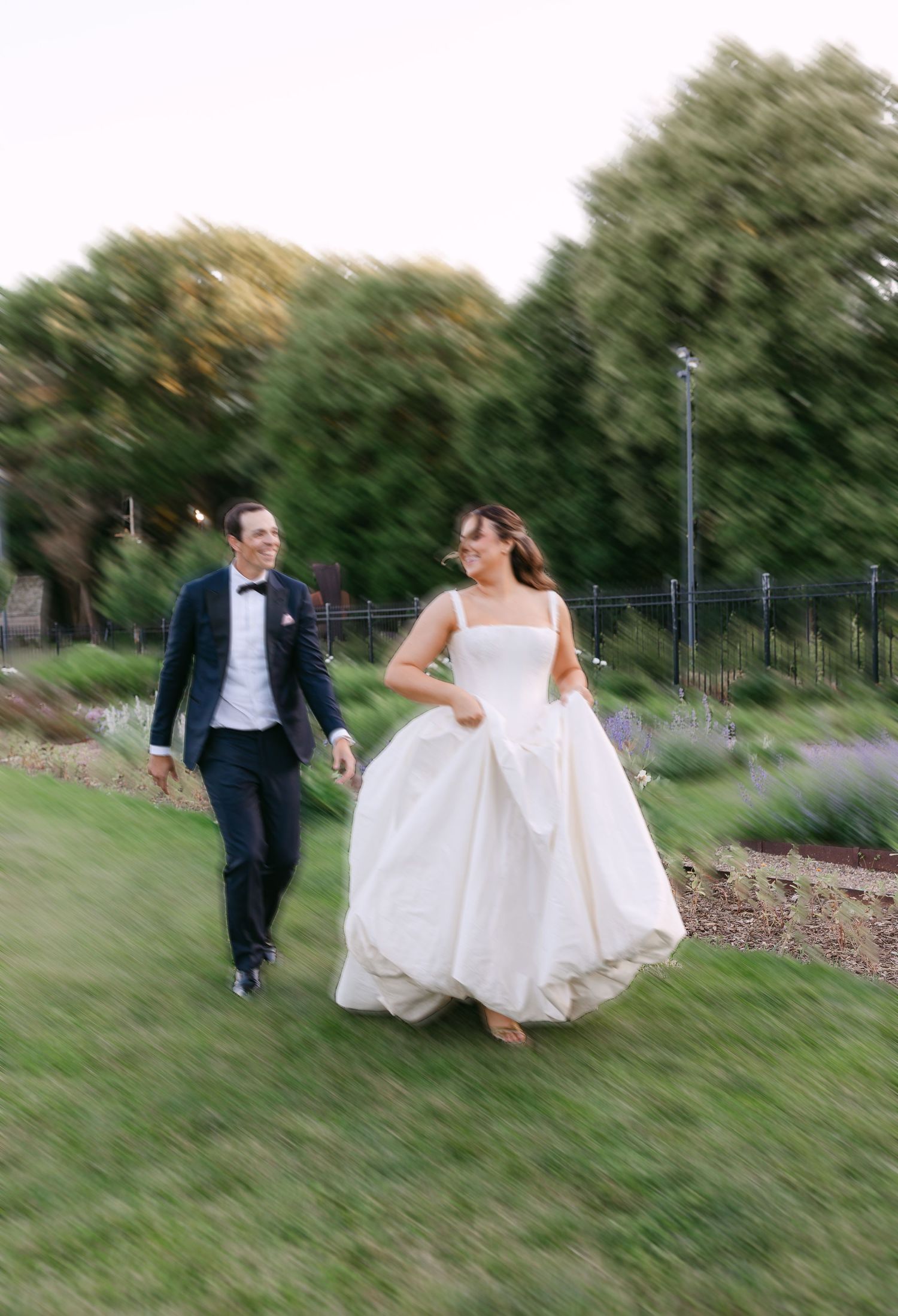 Bride in white ball gown and groom in navy tuxedo running and laughing together across a garden lawn.
