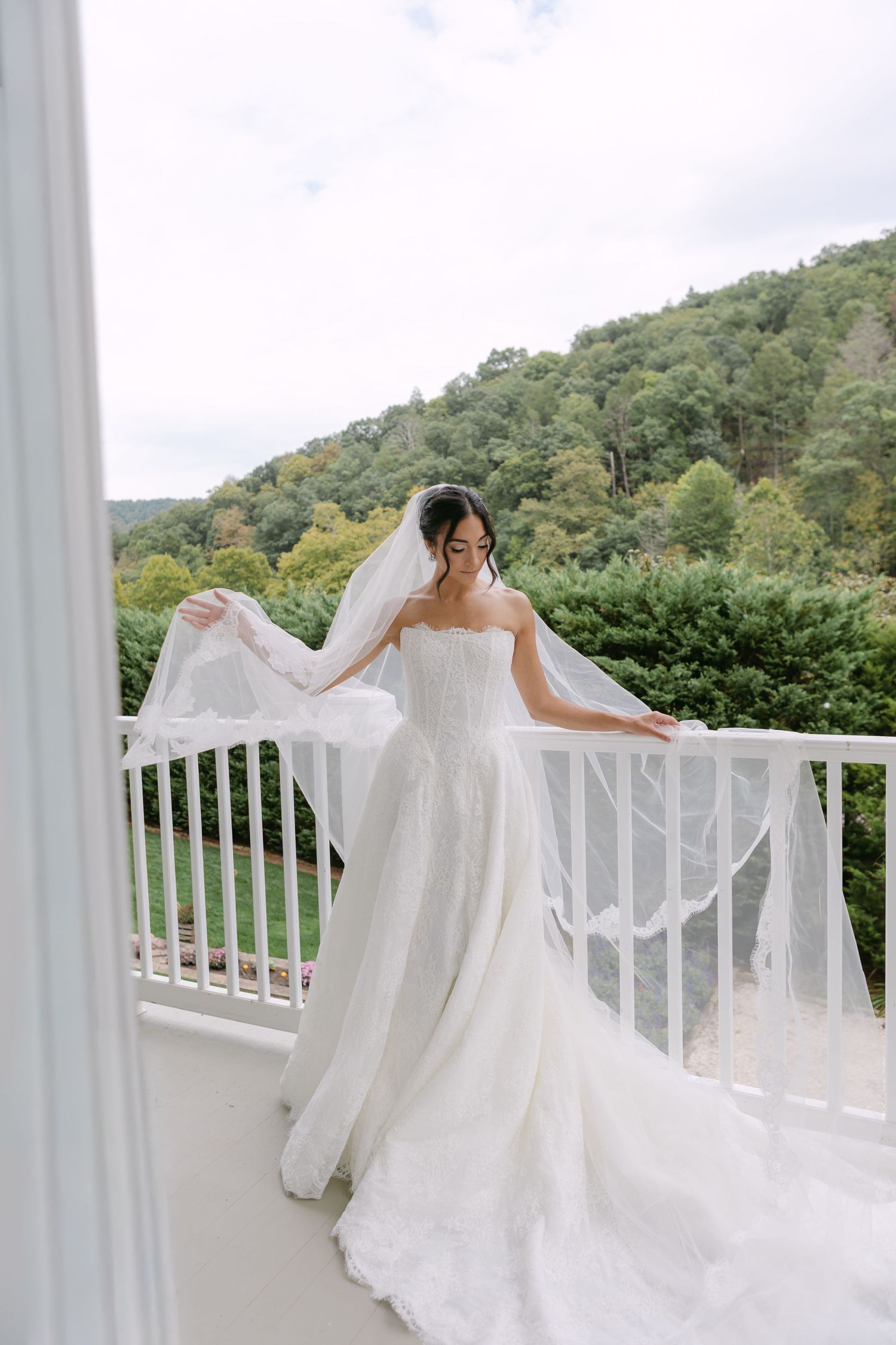 Bride in strapless white ballgown with flowing veil stands on white balcony overlooking lush green mountain landscape.