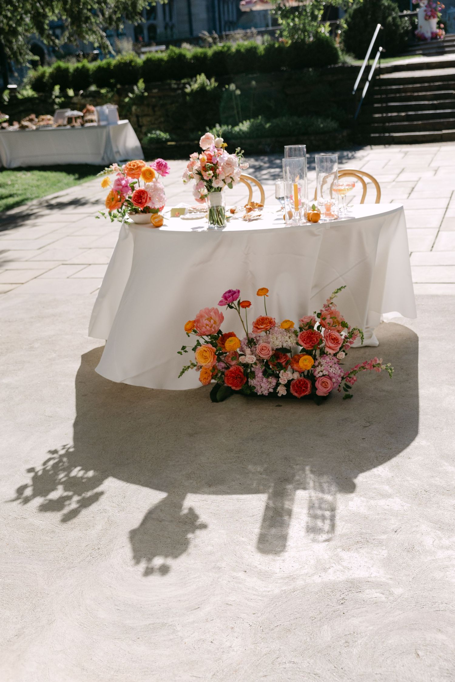 Elegant outdoor wedding sweetheart table with white linen and vibrant pink, orange floral arrangements on sunny patio.