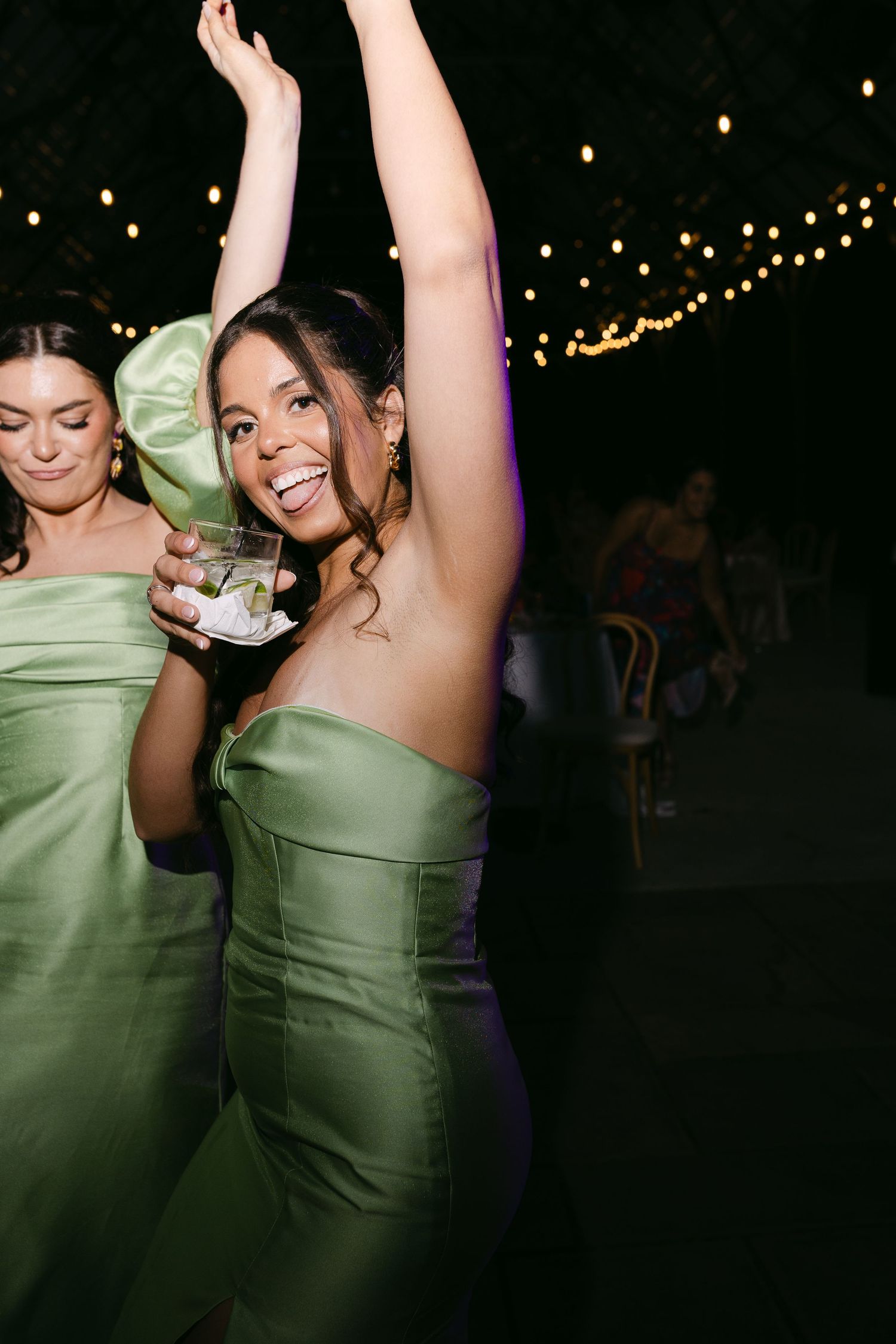 Smiling bridesmaid in green strapless dress dancing with raised arm holding cocktail at wedding reception.