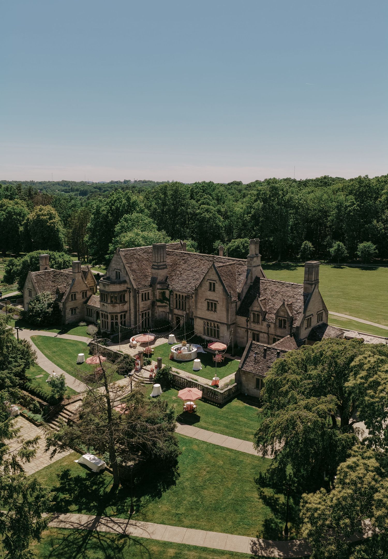 Aerial view of a grand Tudor-style mansion surrounded by lush green trees and manicured lawns with outdoor event setup.