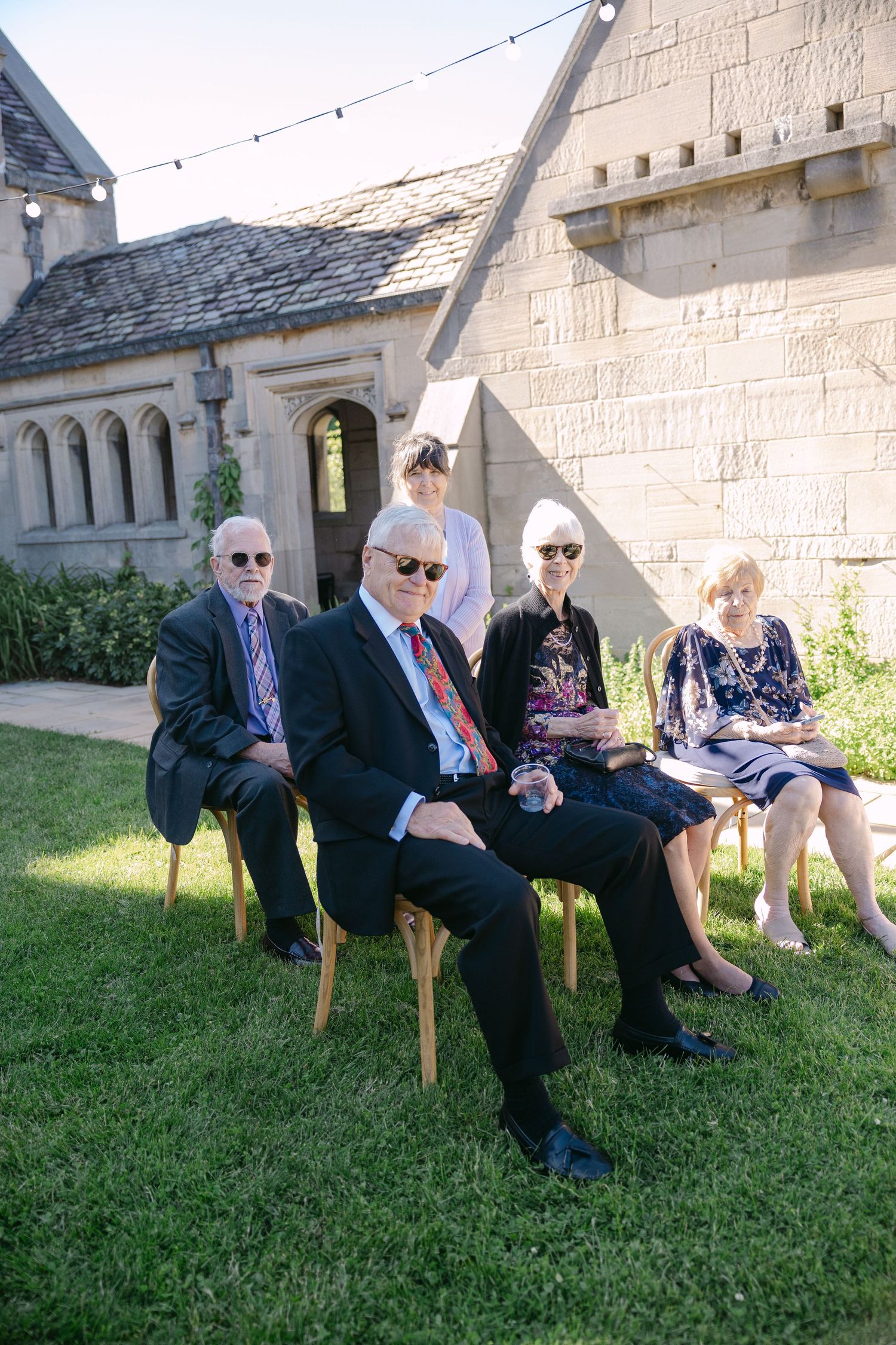 Wedding guests seated on chairs outdoors near stone building with string lights overhead.