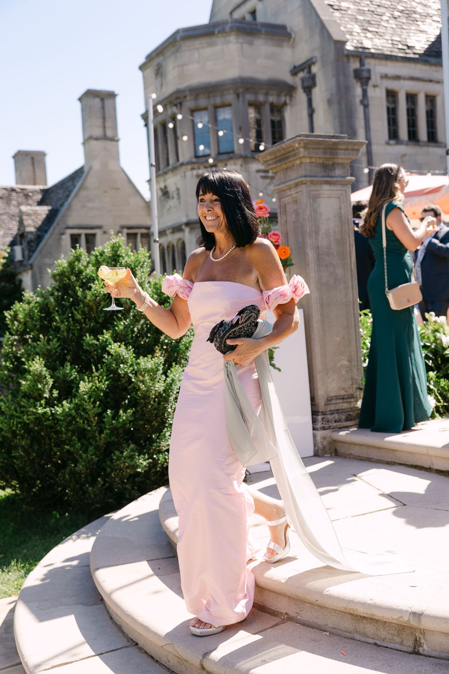A bridesmaid in a pink off-shoulder gown holds champagne glasses outside a stone manor wedding venue.