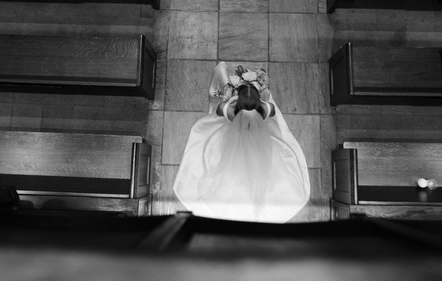 Black and white aerial view of bride in white gown kneeling in prayer between church pews.