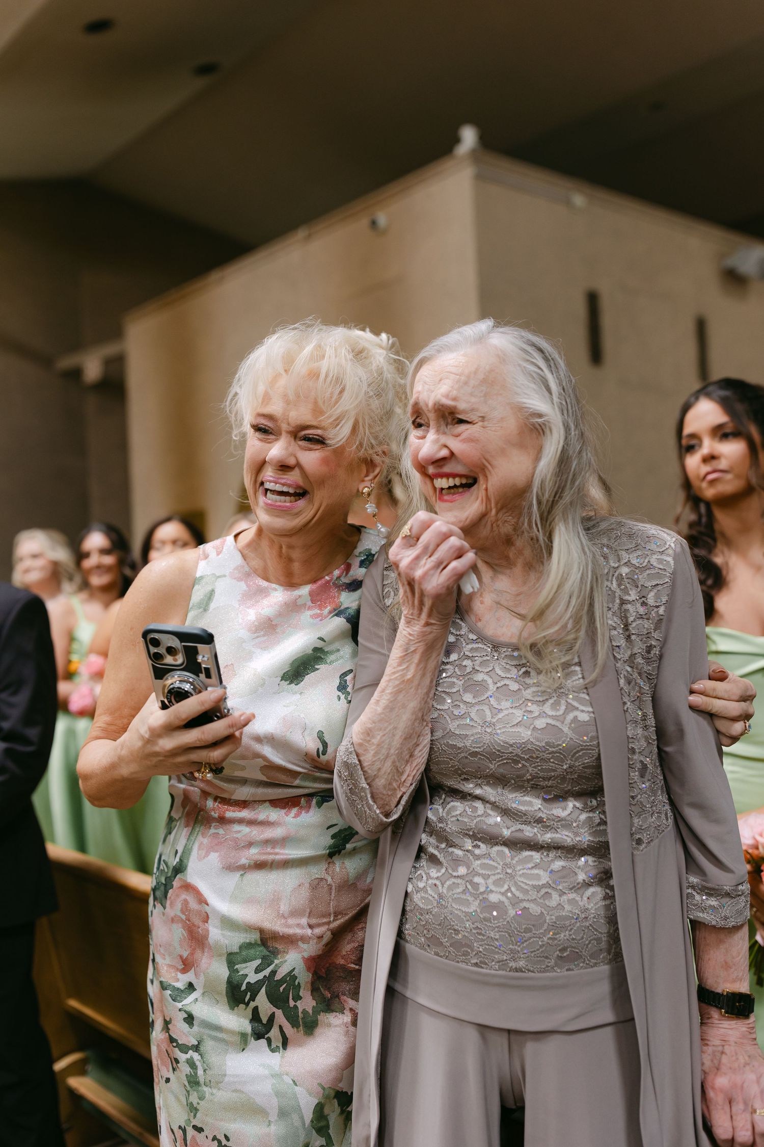 Two elderly women laughing joyfully at a wedding ceremony, one holding a smartphone to share the moment.