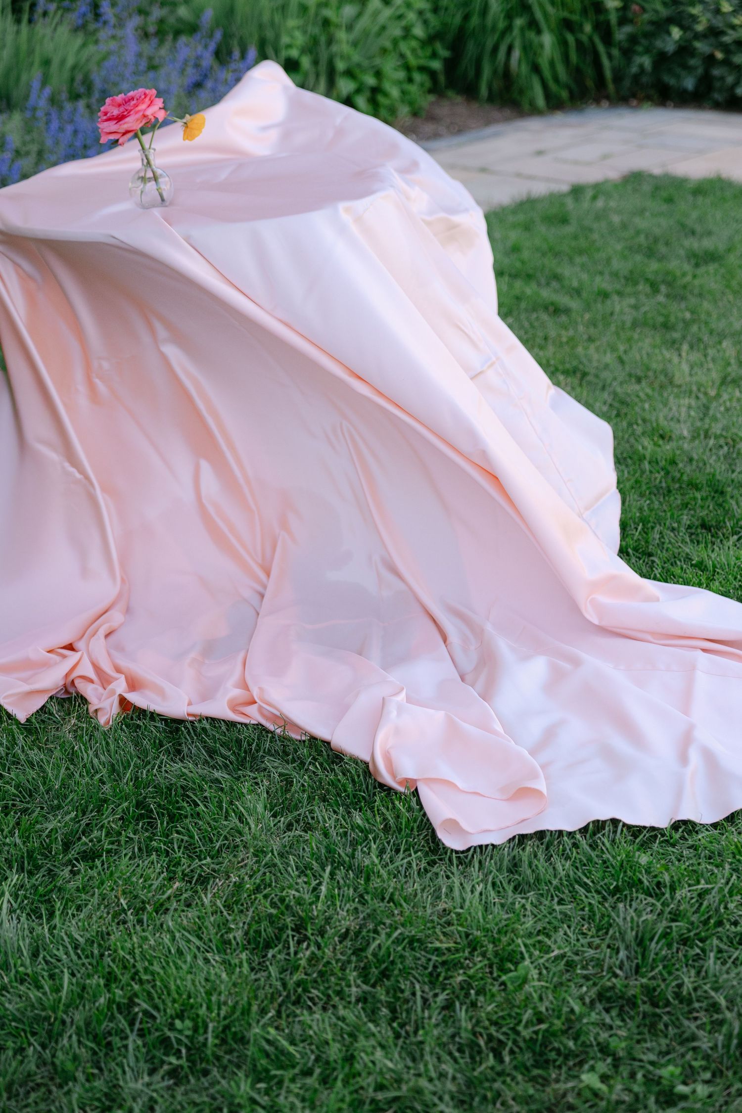 Soft pink draped fabric tablecloth spread across green grass with a single pink rose in a bud vase.