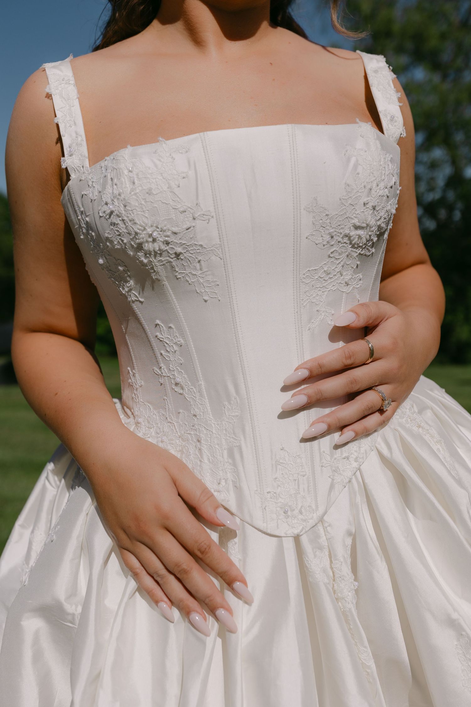 Close-up of bride in white corset wedding gown with lace detailing and hands resting on skirt.