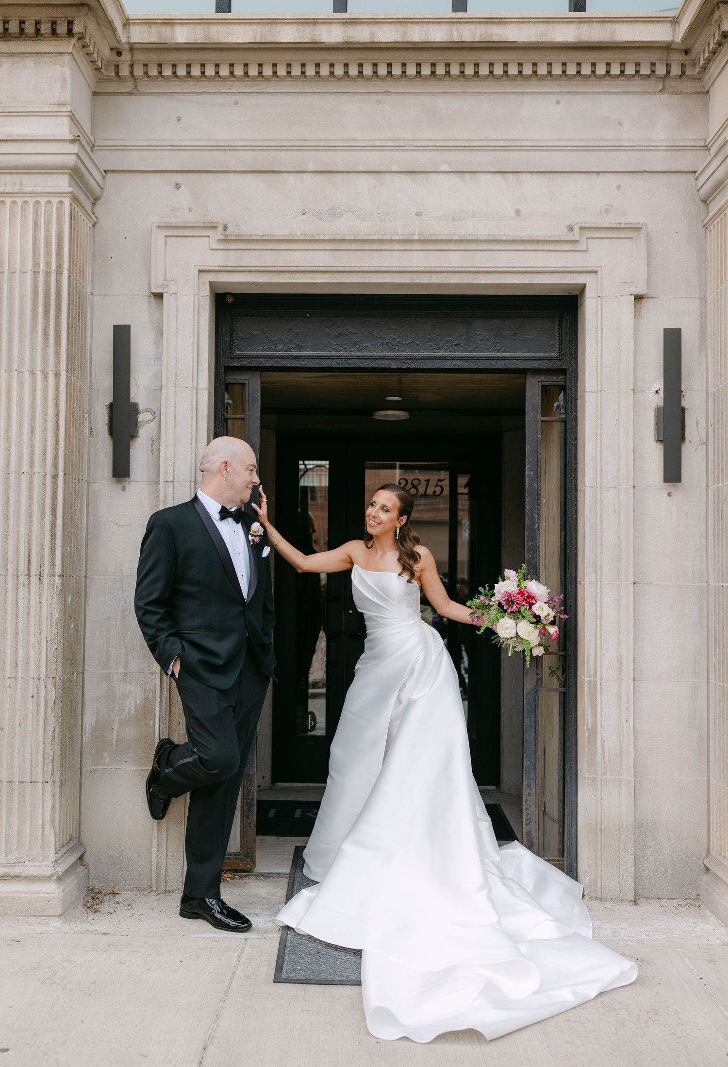 Bride in a white satin gown with groom in tuxedo posing at a grand building entrance.