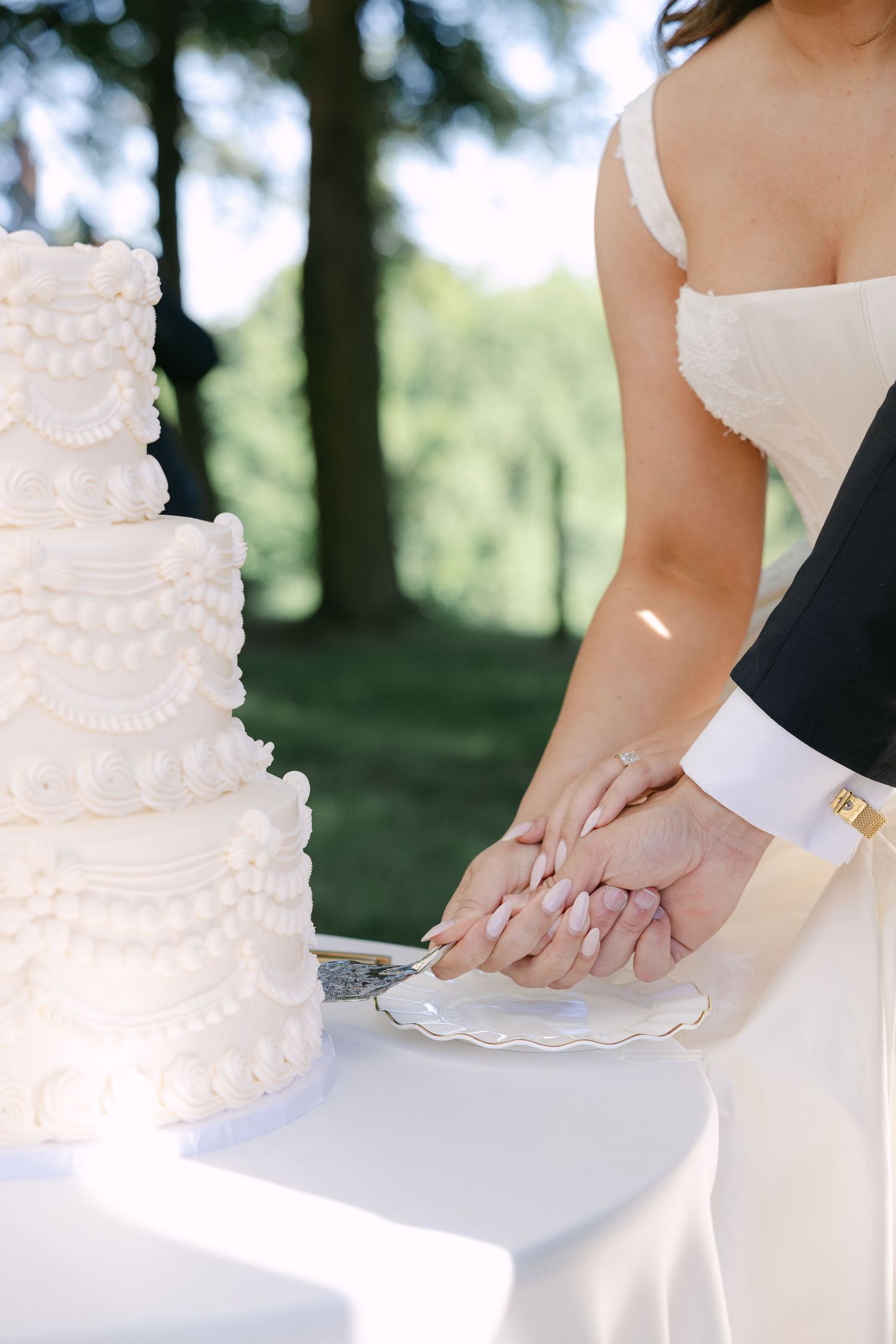 Bride and groom cutting a white four-tier wedding cake with decorative piping outdoors surrounded by trees.