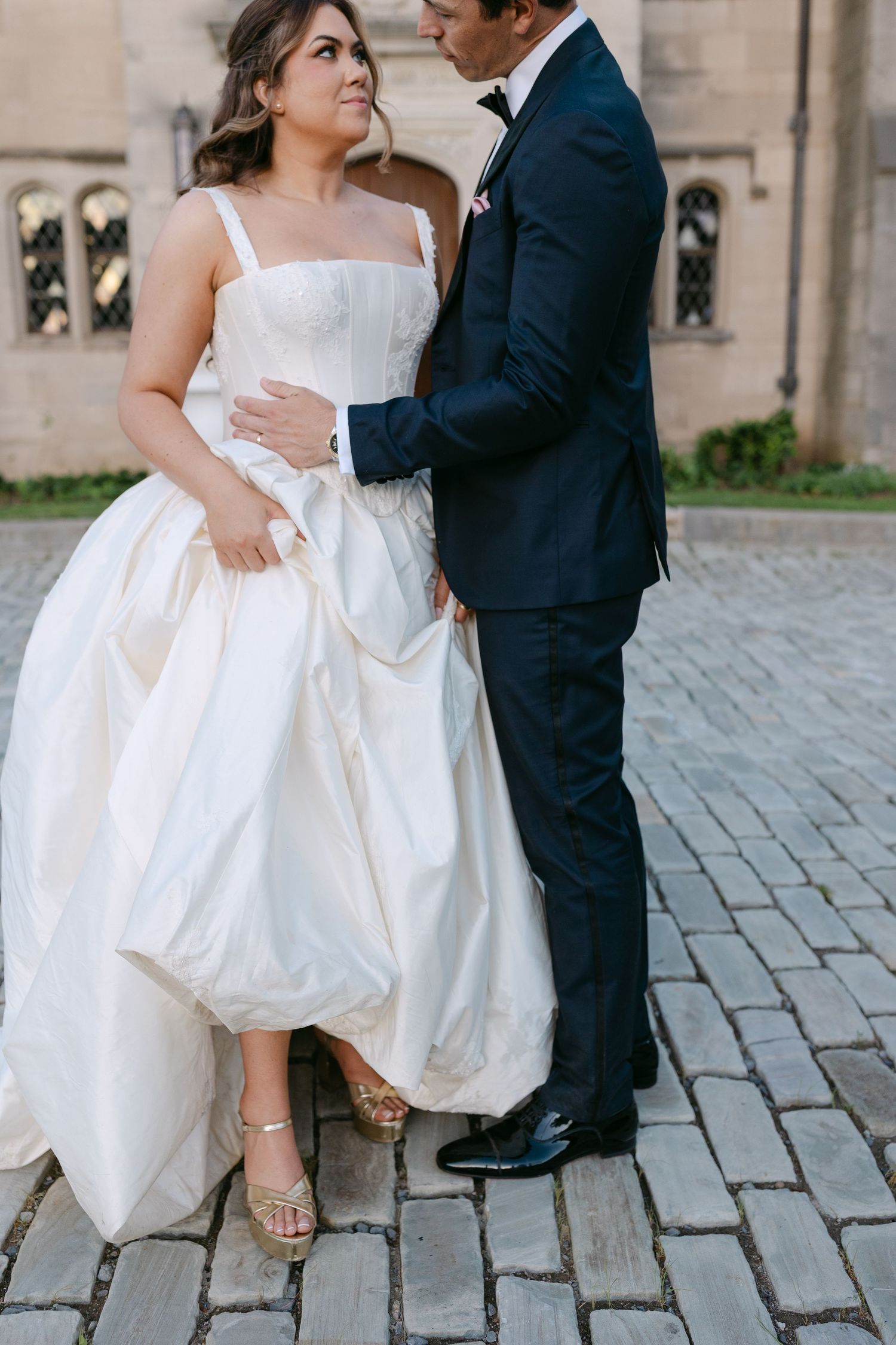 Bride in a white corset ball gown and groom in a navy suit embrace on cobblestone outside a stone church.