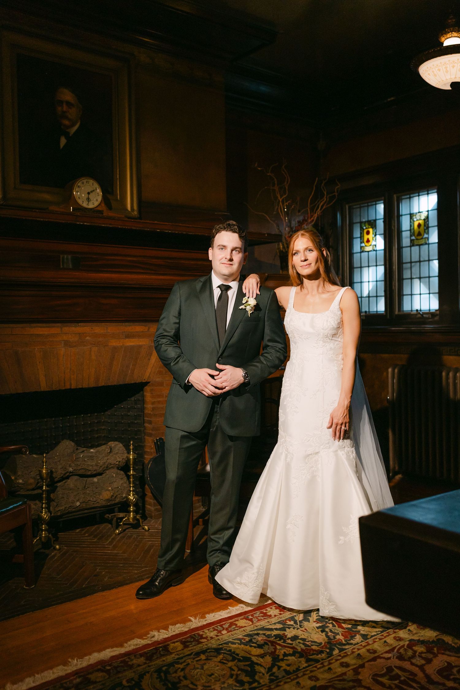 Bride and groom pose together in elegant wood-paneled room with fireplace and stained glass windows.