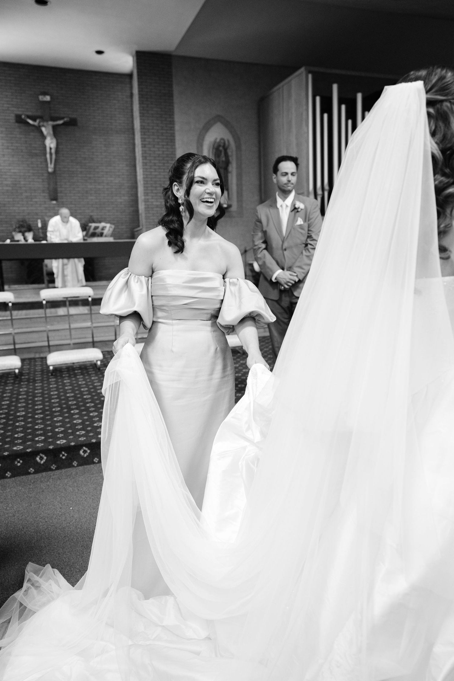 Bride smiling in elegant off-shoulder gown with flowing veil inside a church during wedding ceremony.
