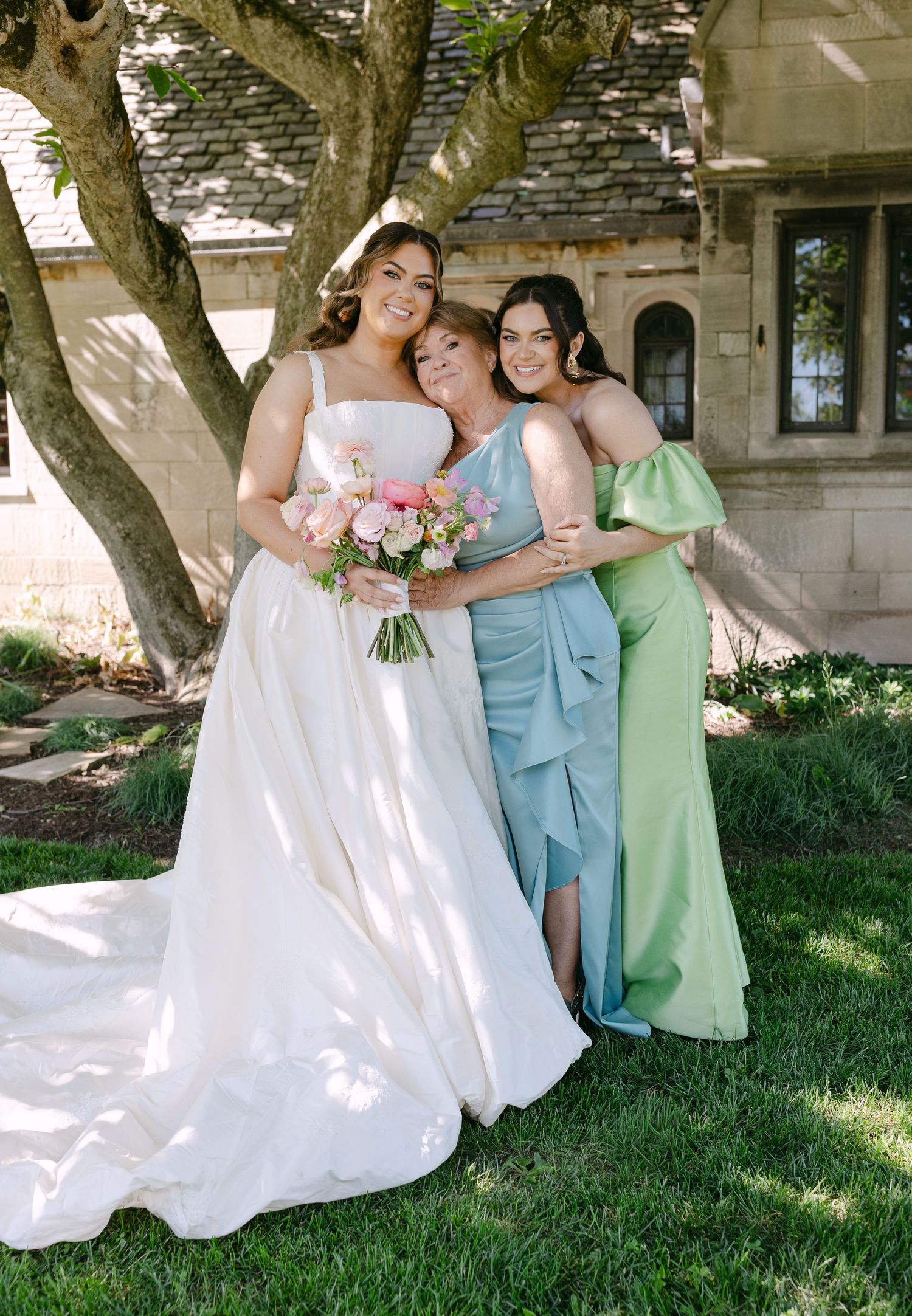 Bride in white ball gown holding pink bouquet poses with two bridesmaids in blue and green dresses outdoors.