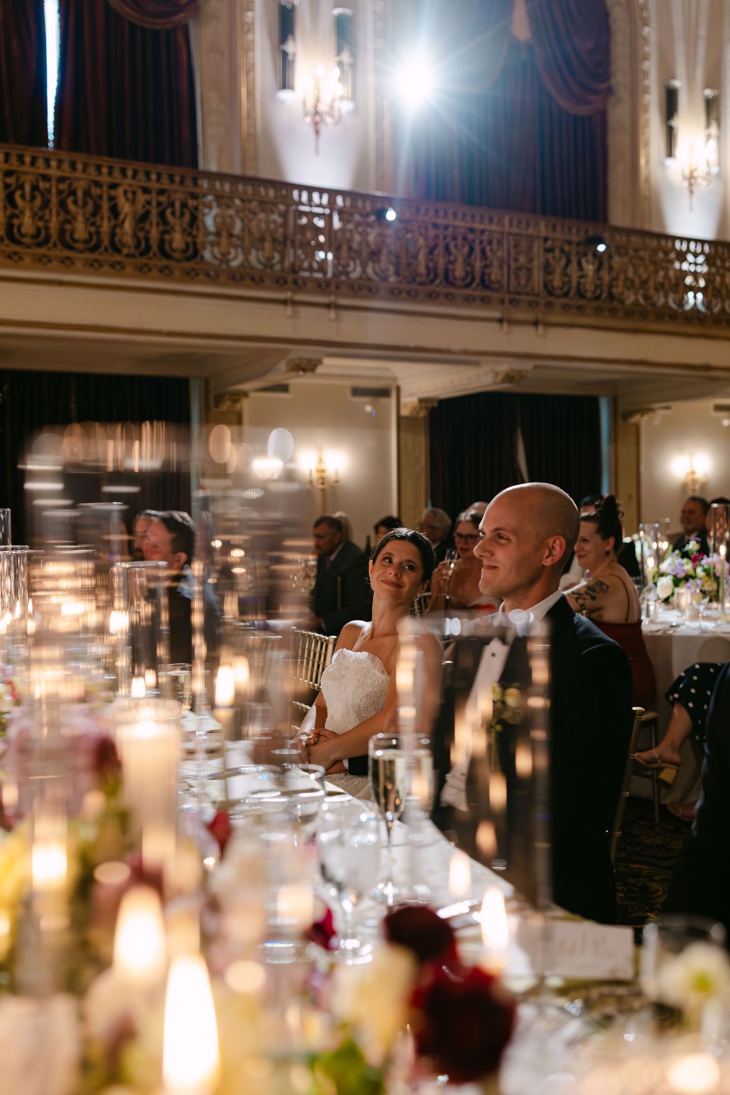 Bride and groom seated at candlelit head table in grand ornate ballroom during elegant wedding reception dinner.