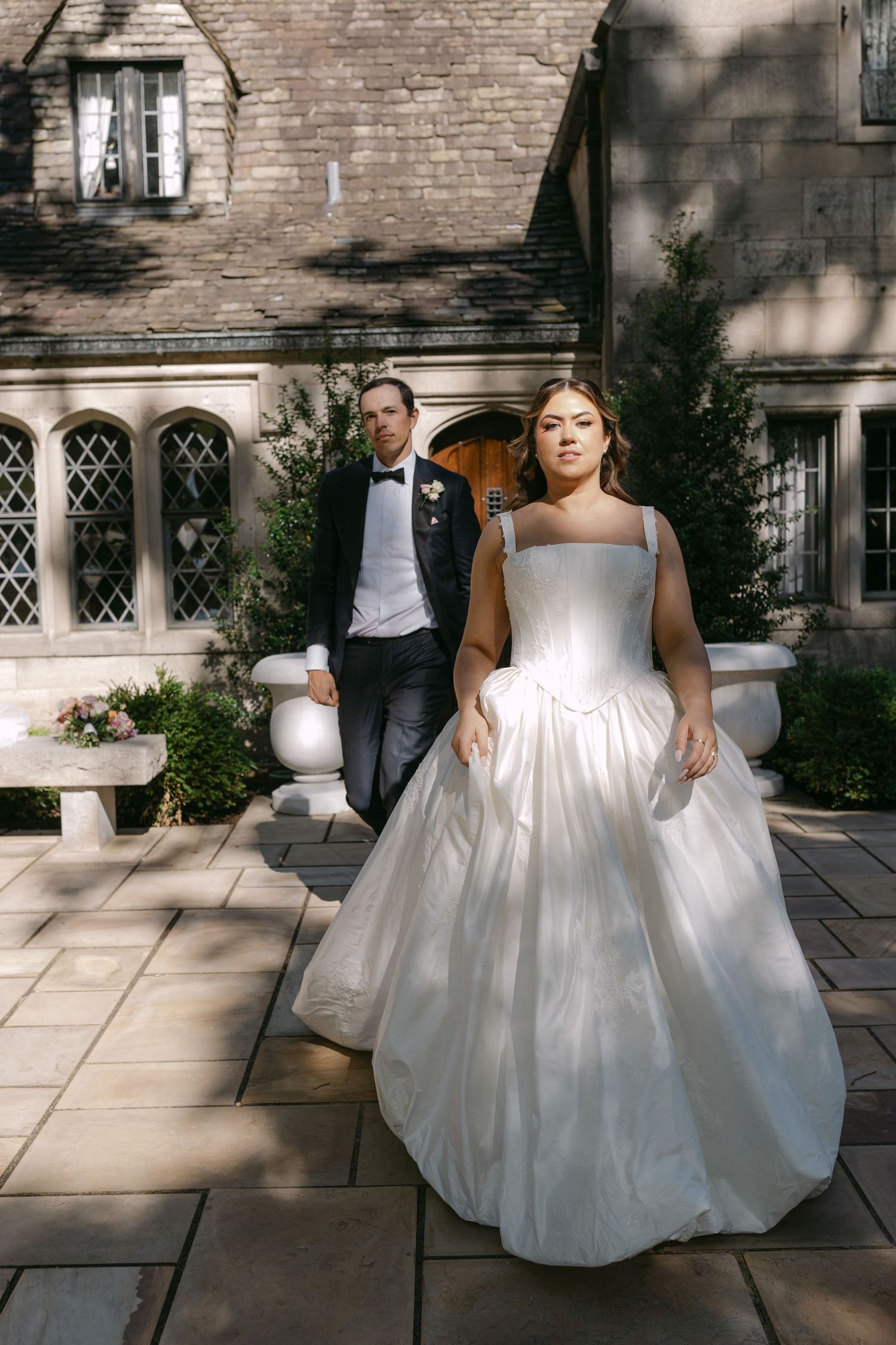 Bride in white square-neck ballgown walks ahead of groom in black tuxedo at stone manor wedding venue.