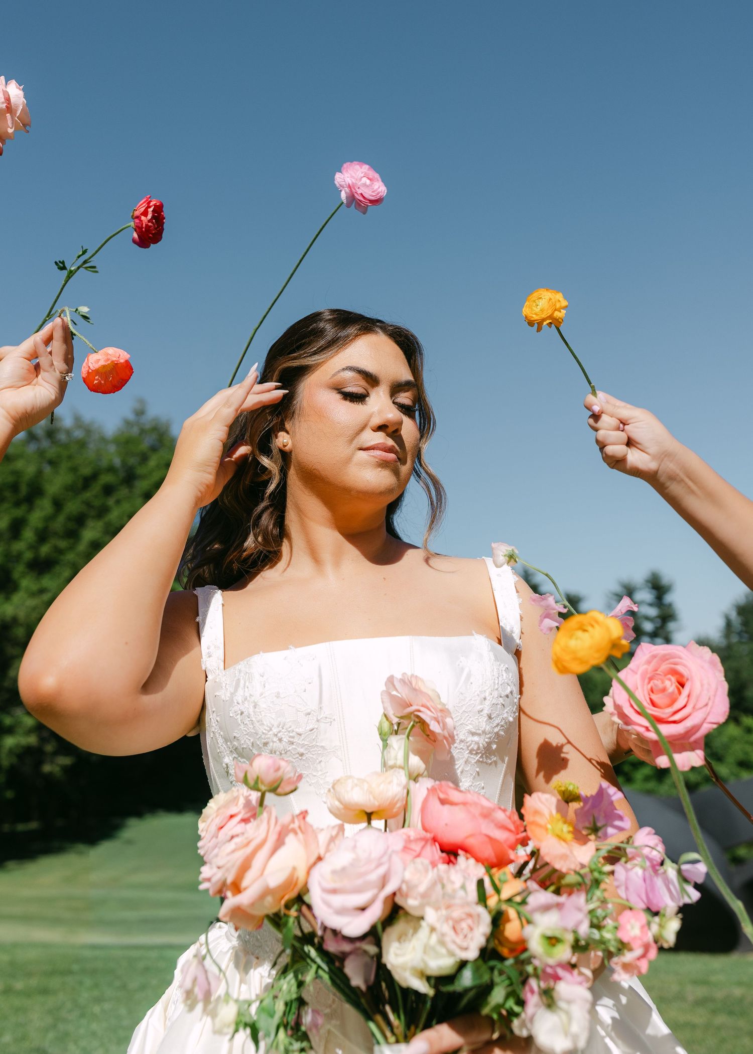 Bride in white gown holds colorful bouquet as hands offer vibrant ranunculus flowers around her.