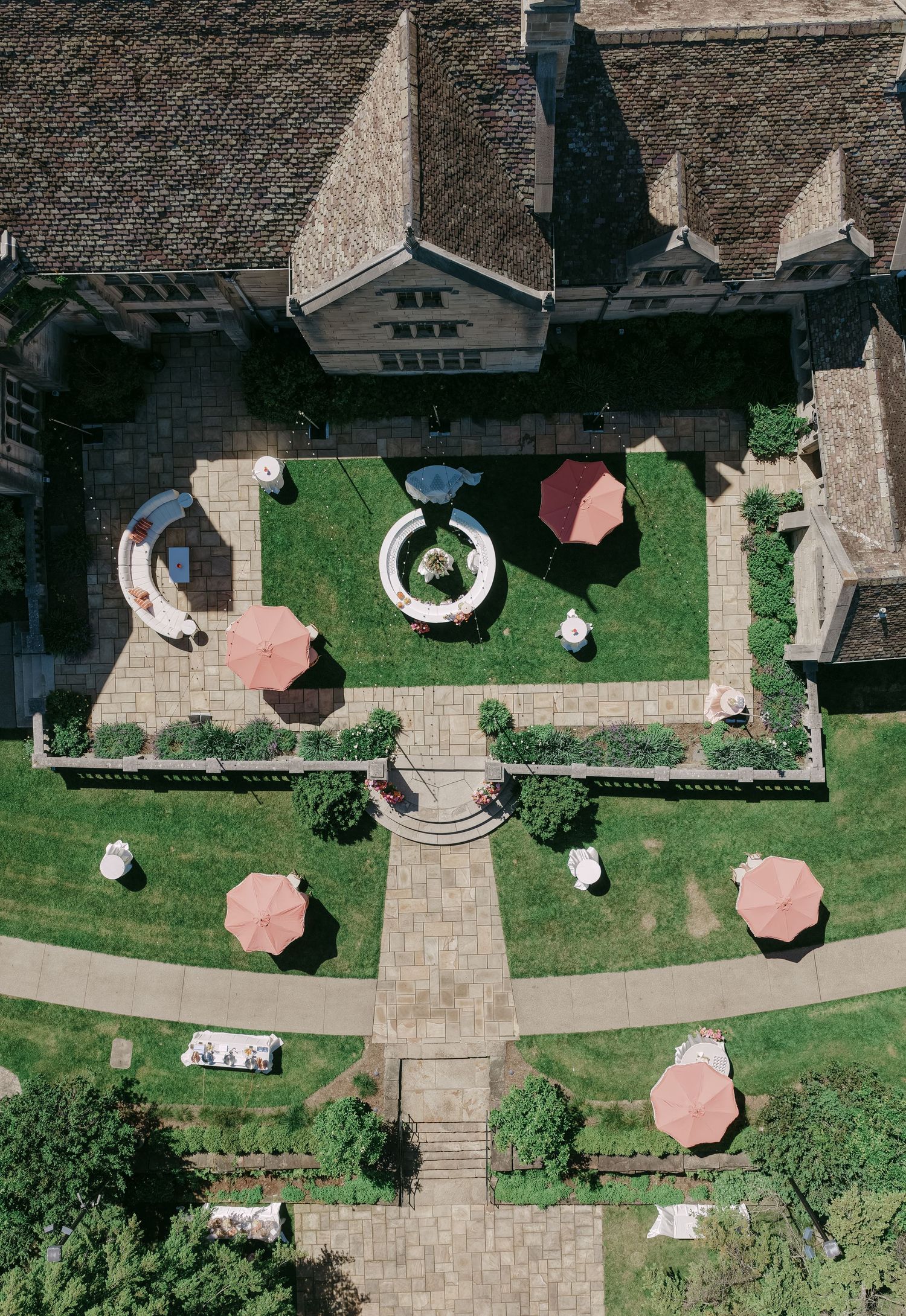 Aerial view of elegant estate courtyard with manicured gardens, fountain, and pink patio umbrellas.