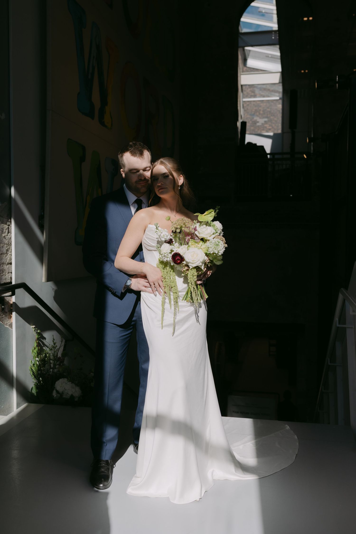 Bride and groom posing in dramatic light and shadow in an archway, bride holding lush white and burgundy wedding bouquet.