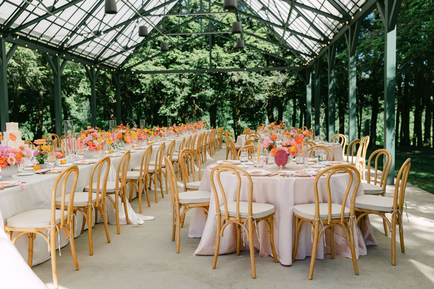Spacious greenhouse wedding reception with round tables, gold chairs, blush linens, and colorful centerpieces.
