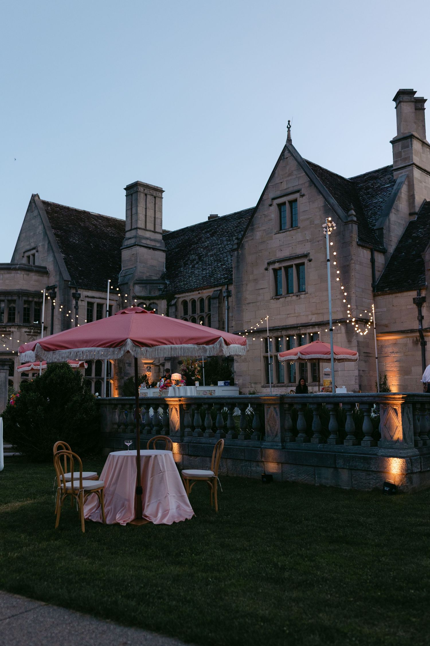 Stone manor wedding venue at dusk with pink umbrellas, string lights, and elegant outdoor seating.