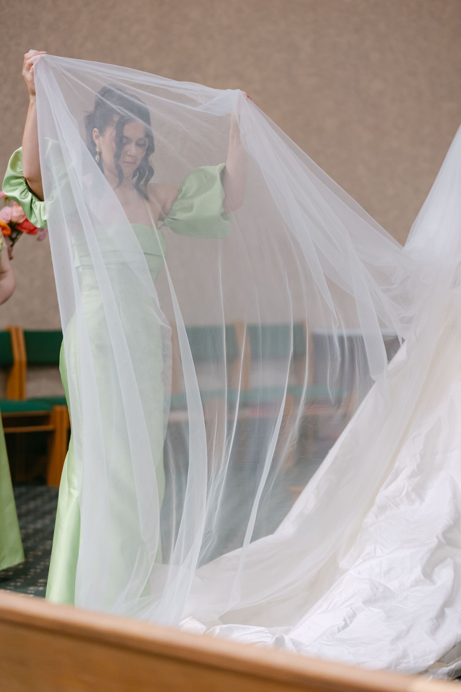 Bridesmaid in mint green dress holds up a flowing white bridal veil during wedding preparations indoors.