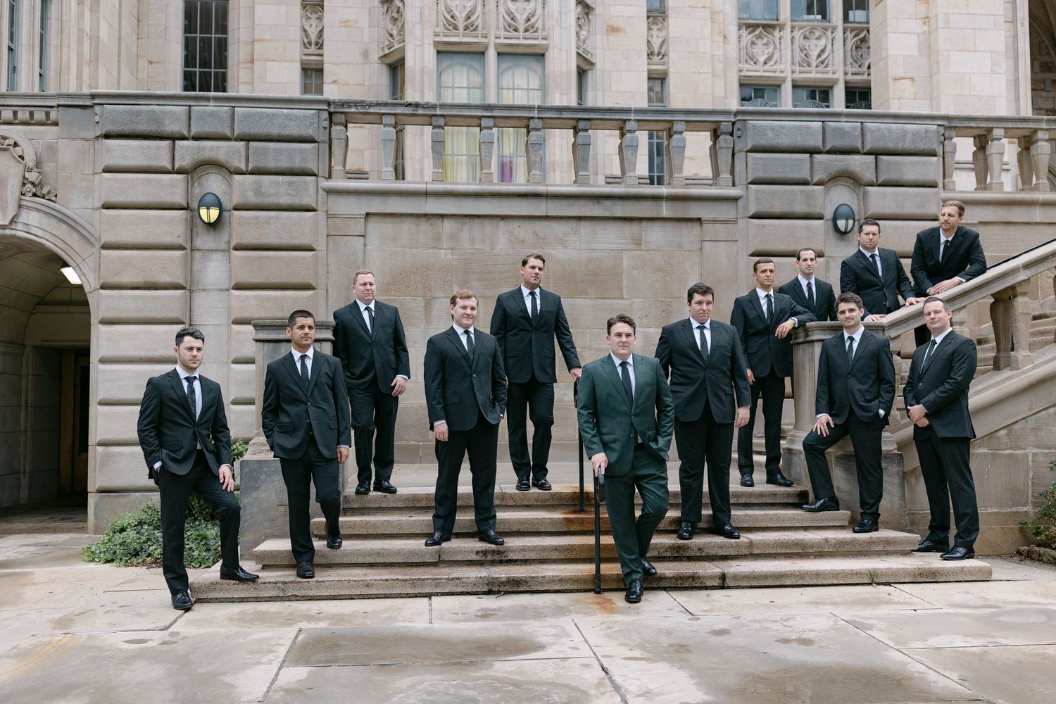 Groomsmen in dark suits pose on stone steps outside a grand classical wedding venue.