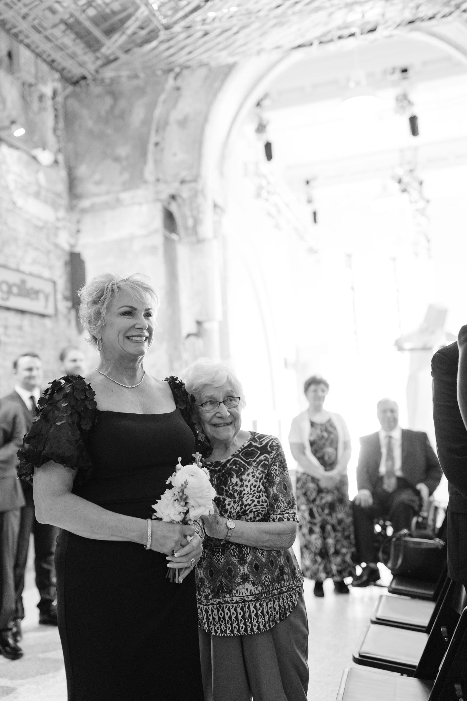 Black and white photo of two women walking down the aisle at a wedding ceremony in an ornate venue.