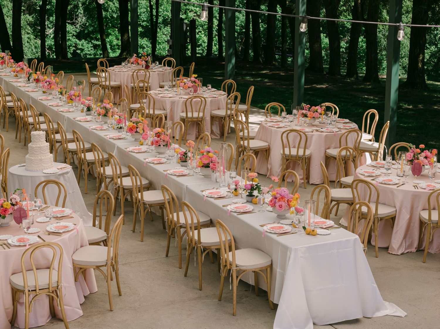 Long and round tables with gold chairs, pink linens, and colorful floral centerpieces at an outdoor wedding reception.