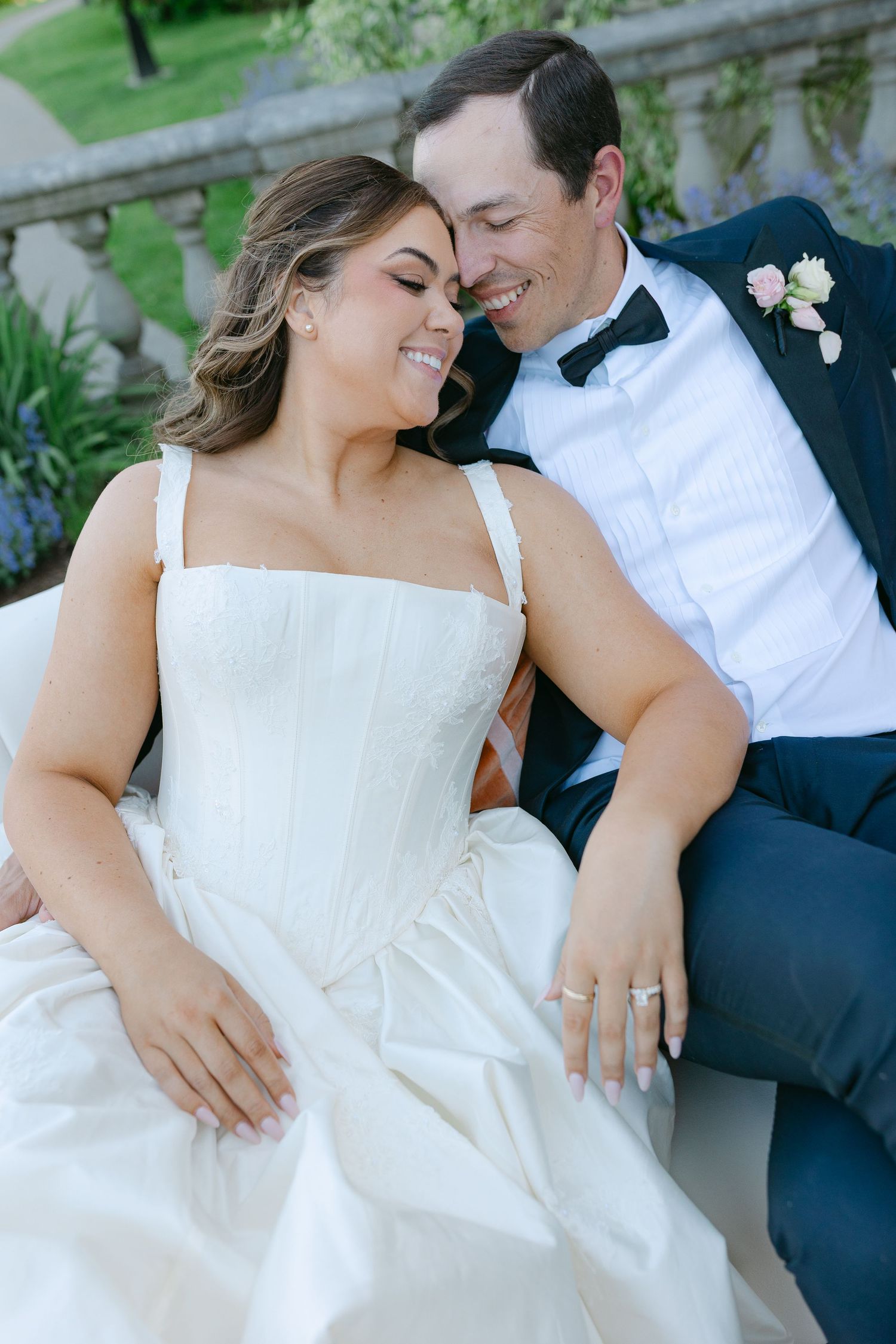 A smiling bride in a white corset gown and groom in navy tuxedo share a tender moment seated outdoors at their wedding.