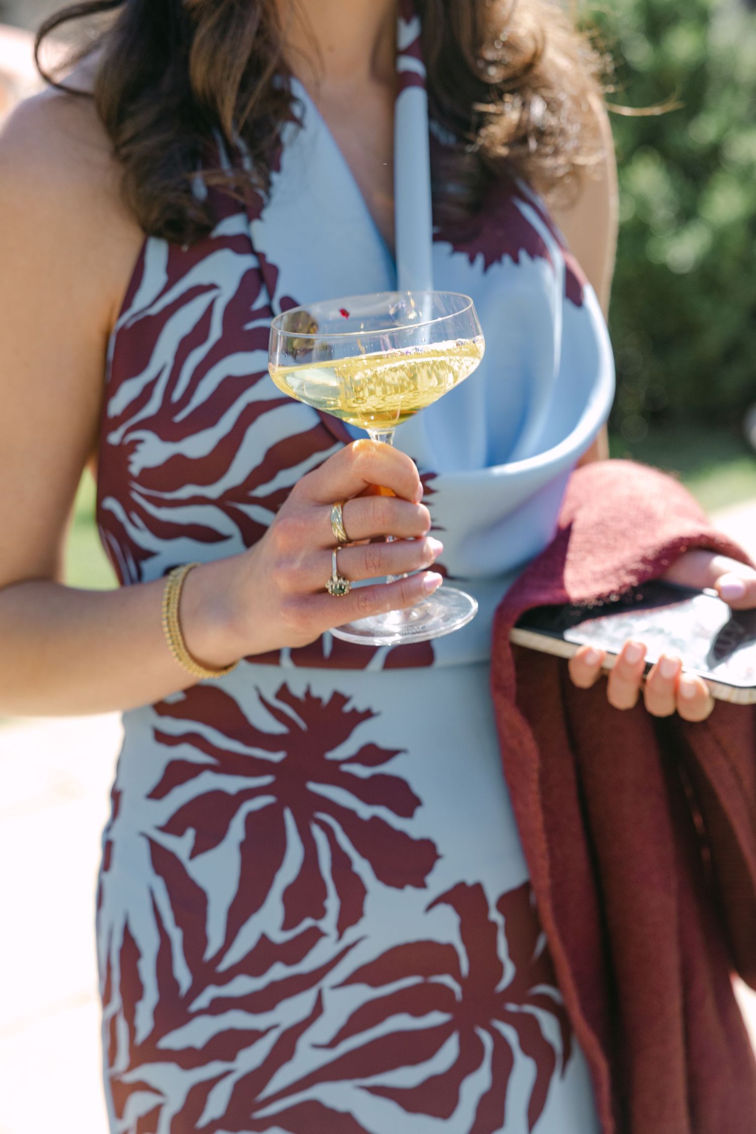 Wedding guest in blue and burgundy floral dress holding champagne coupe glass at outdoor reception.