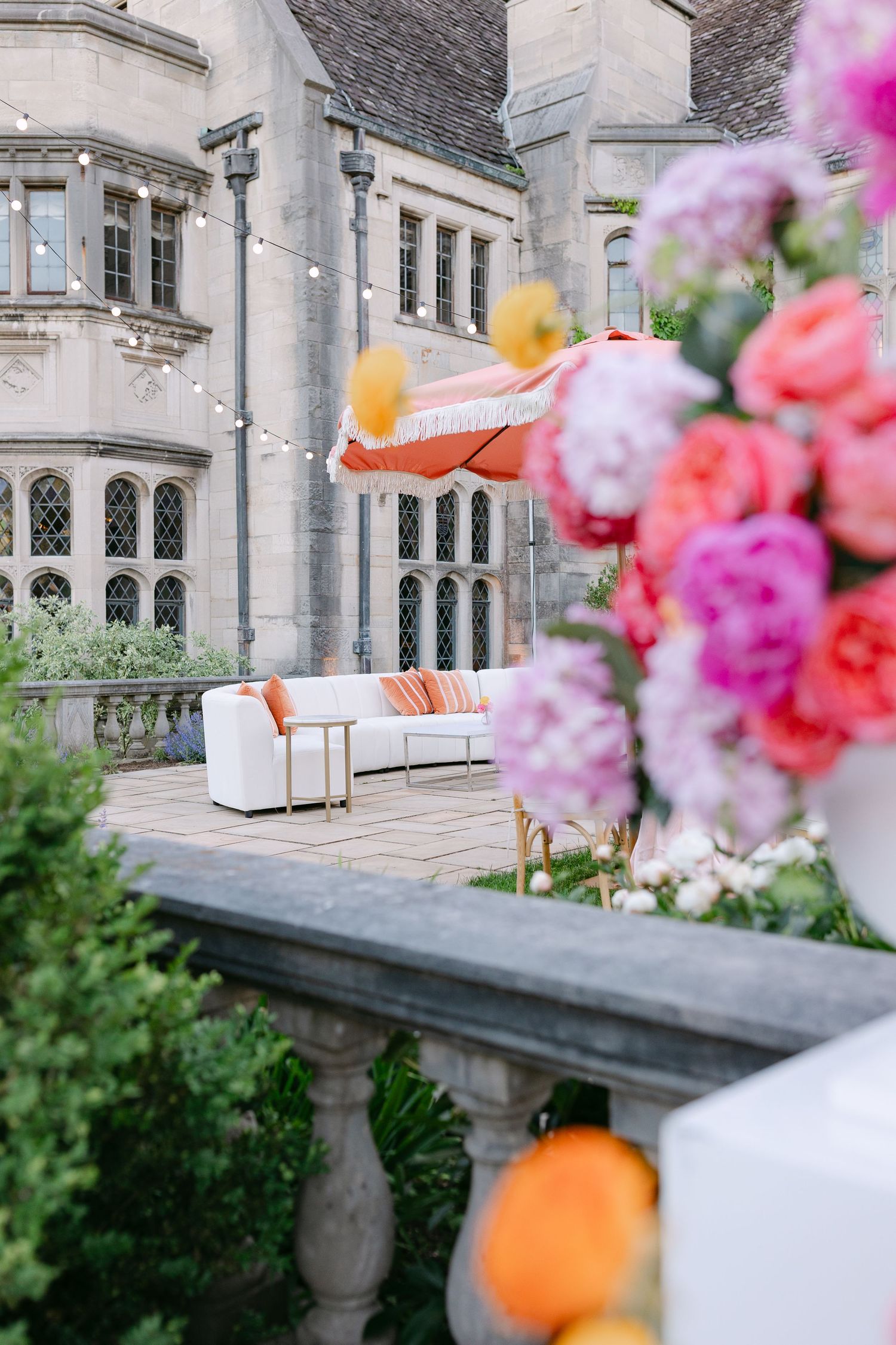 Elegant outdoor wedding lounge with white sofas, orange umbrella, and vibrant pink and coral floral arrangements.